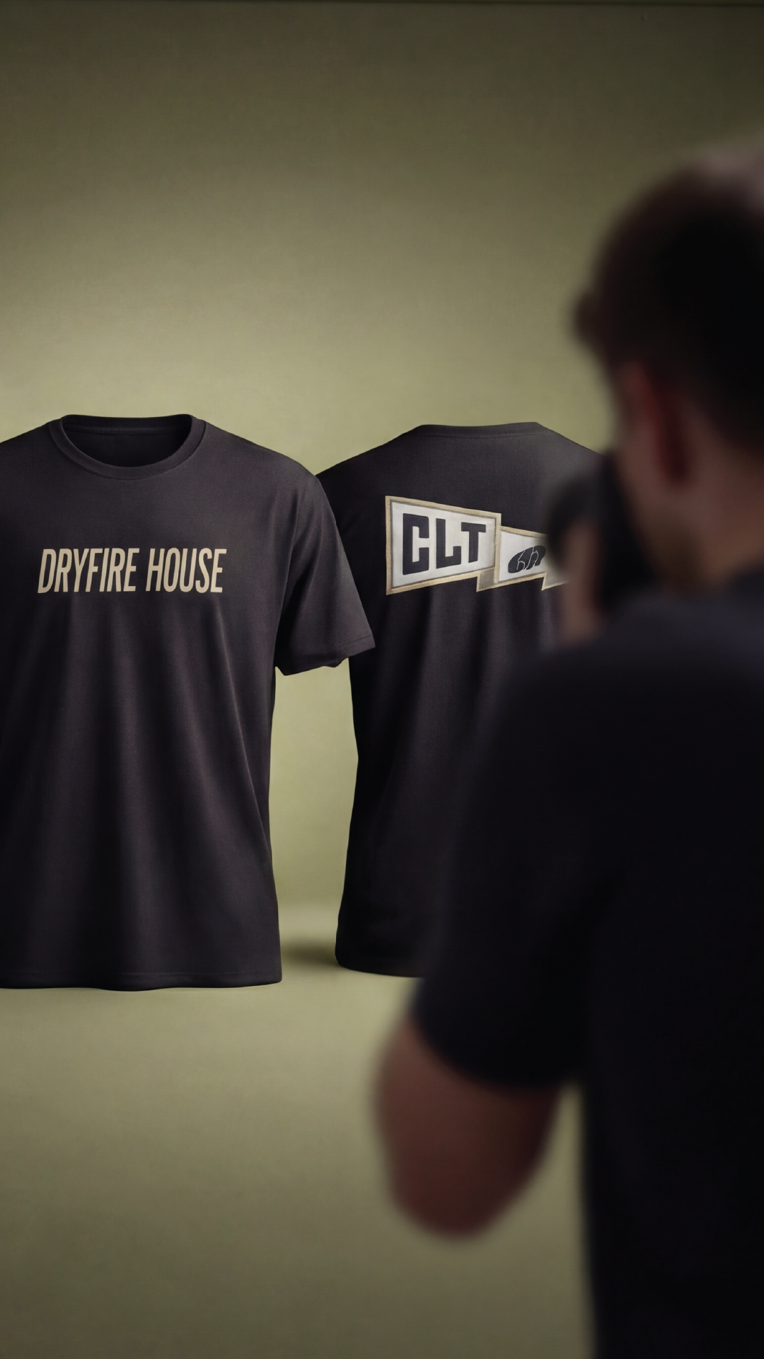 Photo of two black DRYFIRE HOUSE T-shirts hanging on display, one with 'DRYFIRE HOUSE' written on it and the other with 'CLT' branding, with a person in foreground taking a photo.