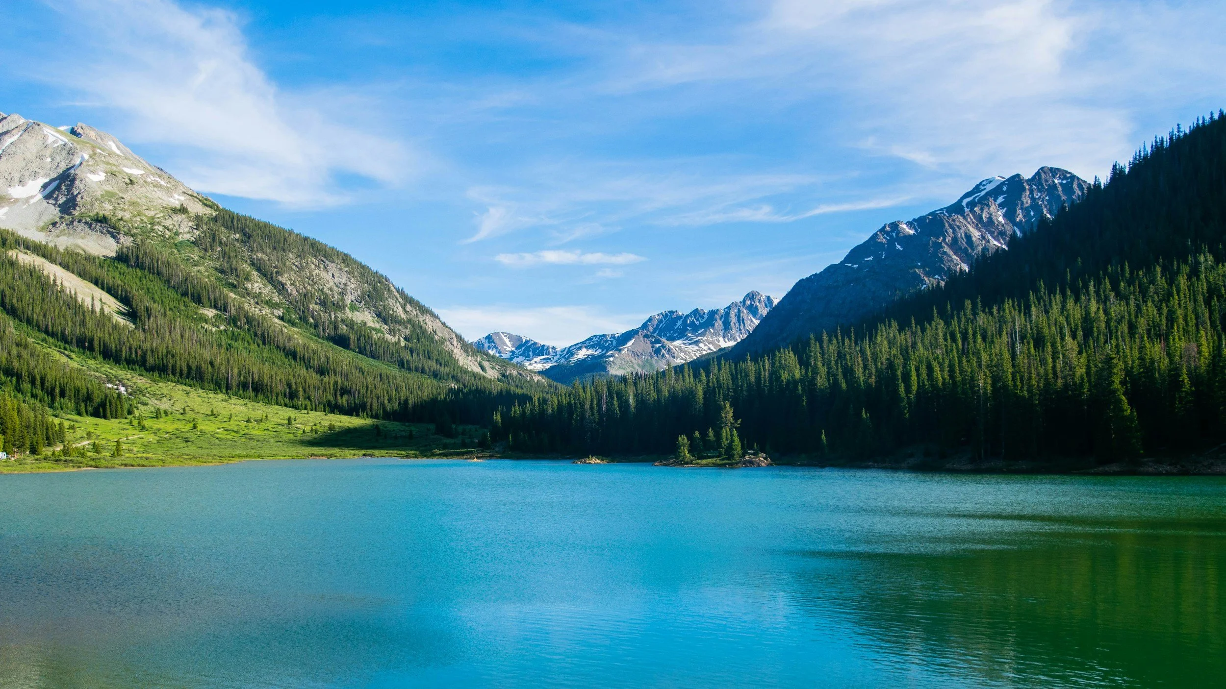 Scenic landscape featuring a calm blue lake surrounded by green forested mountains with snow-capped peaks, under a partly cloudy sky.