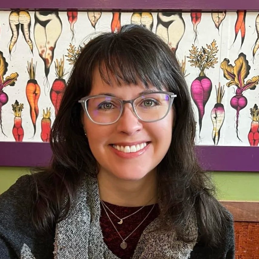Portrait of a woman with glasses smiling, with a colorful vegetable-themed painting of carrots in the background.