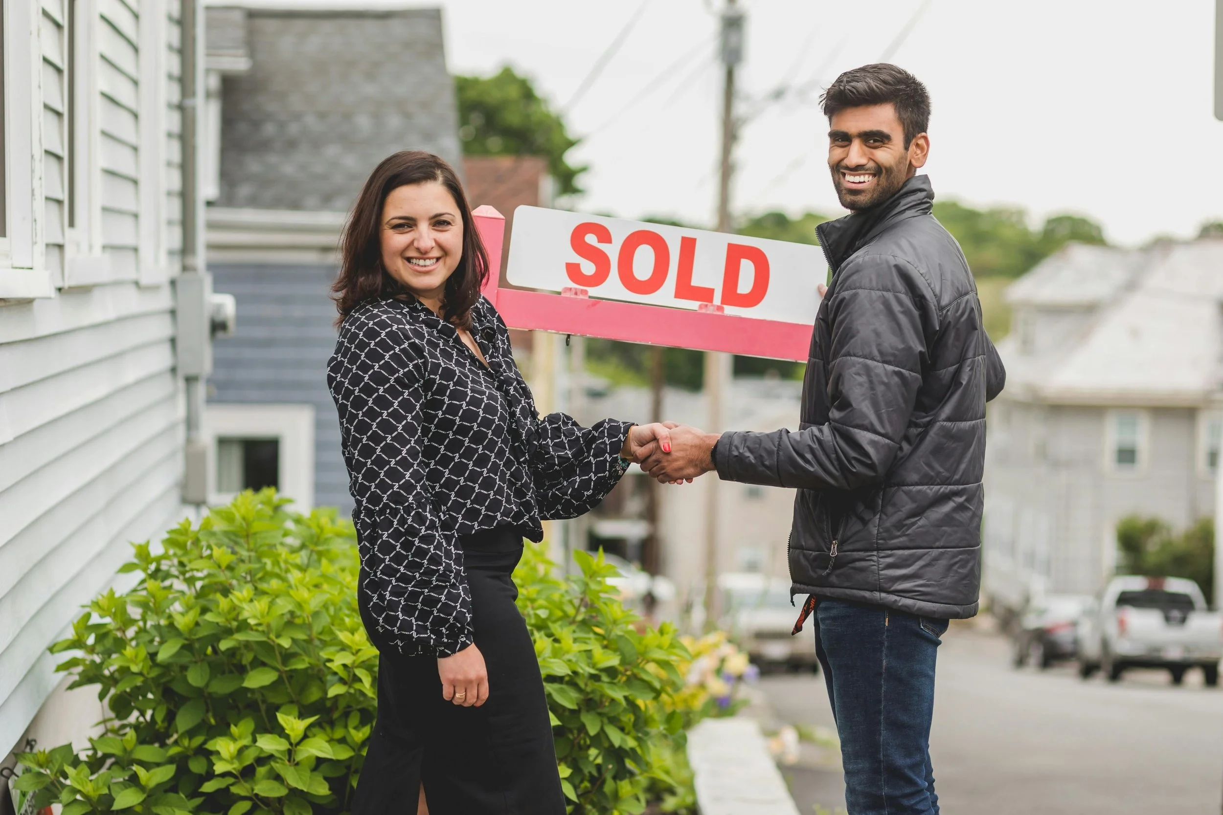 A man and woman shake hands in front of a house in Colorado, smiling, with a 'SOLD' sign between them, indicating a successful home sale.