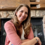 A woman with long brown hair smiling, sitting near a fireplace with a stone surround.
