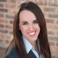 A woman with long dark hair smiling, wearing a light blue blouse and dark sweater, standing in front of a brick wall.