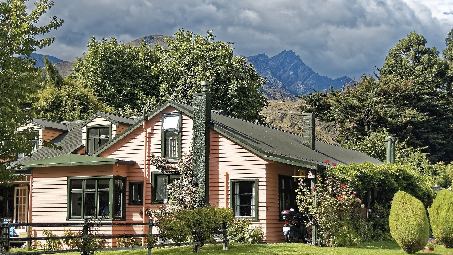 A light pink house in Colorado with black window frames and a green roof, surrounded by trees, bushes, and flowers, with a mountain range in the background and cloudy sky above.