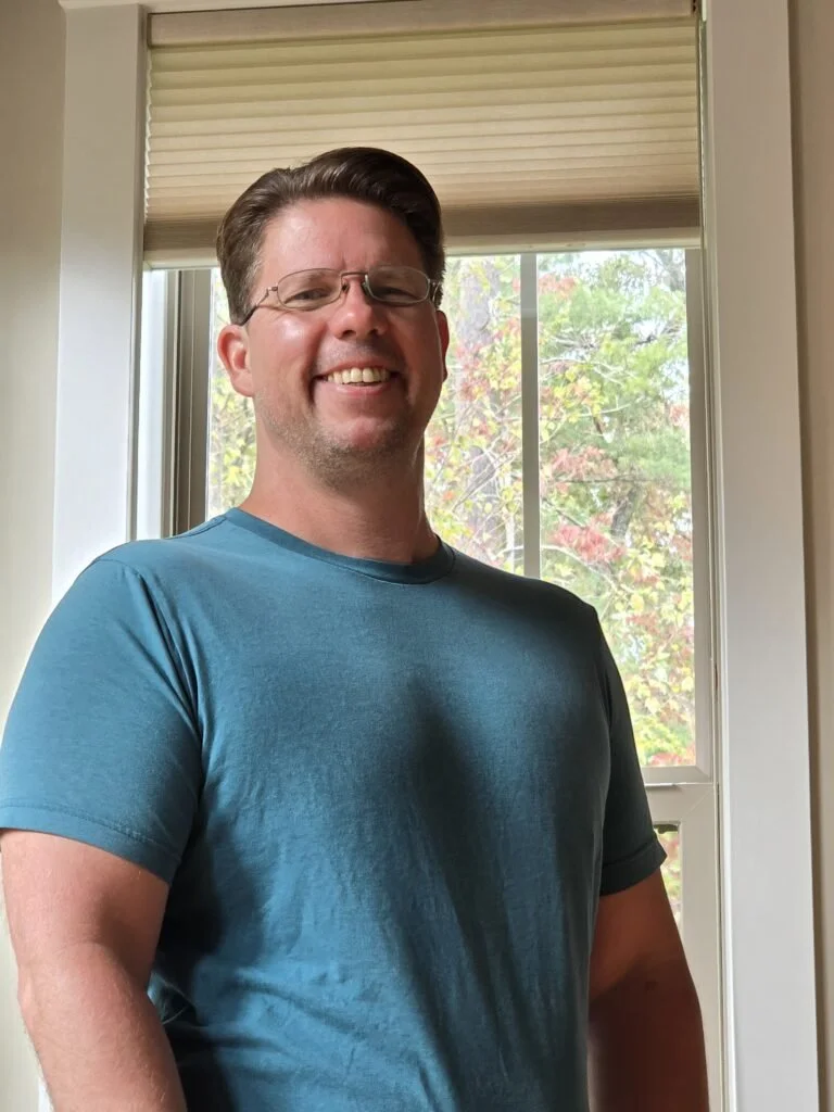 A man with glasses and brown hair smiling, wearing a teal t-shirt, standing indoors in front of a window with trees outside.