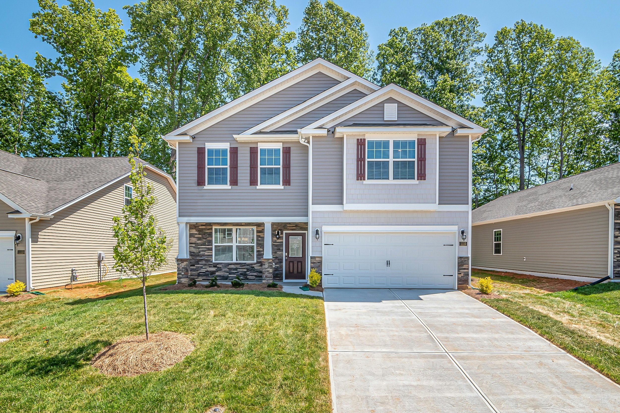A two-story suburban house with gray siding, white trim, and a stone accent on the lower front. It has three windows with maroon shutters on the upper level and a large window with similar shutters on the front. The house has a white garage door and 