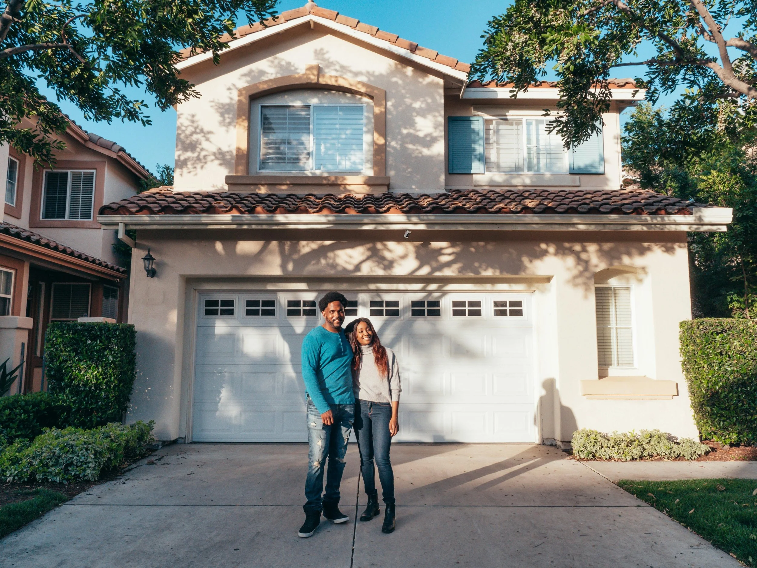 A man and a woman standing in front of a white house in Colorado with a garage door, surrounded by green trees and bushes. The man is wearing a teal shirt and jeans, and the woman is wearing a light-colored top and boots.