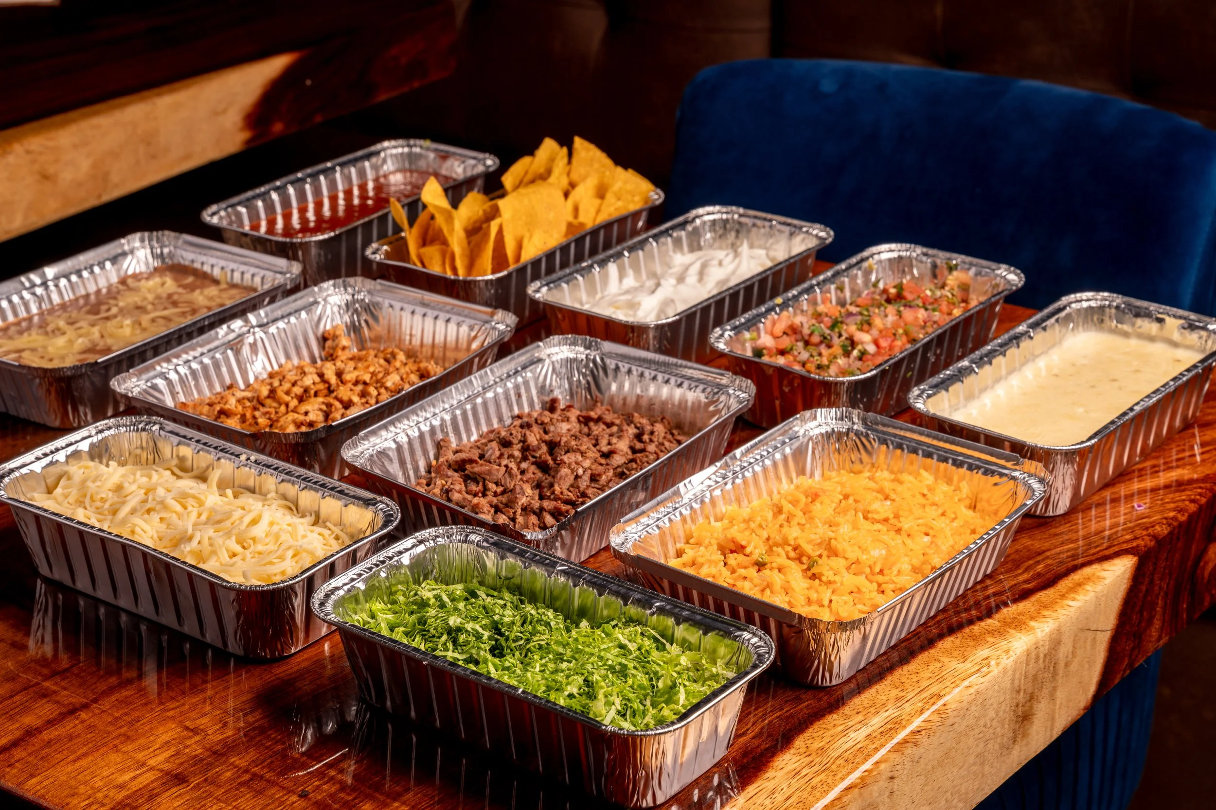 Aluminum containers filled with various Mexican dishes and toppings on a wooden table, including shredded cheese, cooked ground meat, shredded lettuce, salsa, sour cream, and diced tomatoes.
