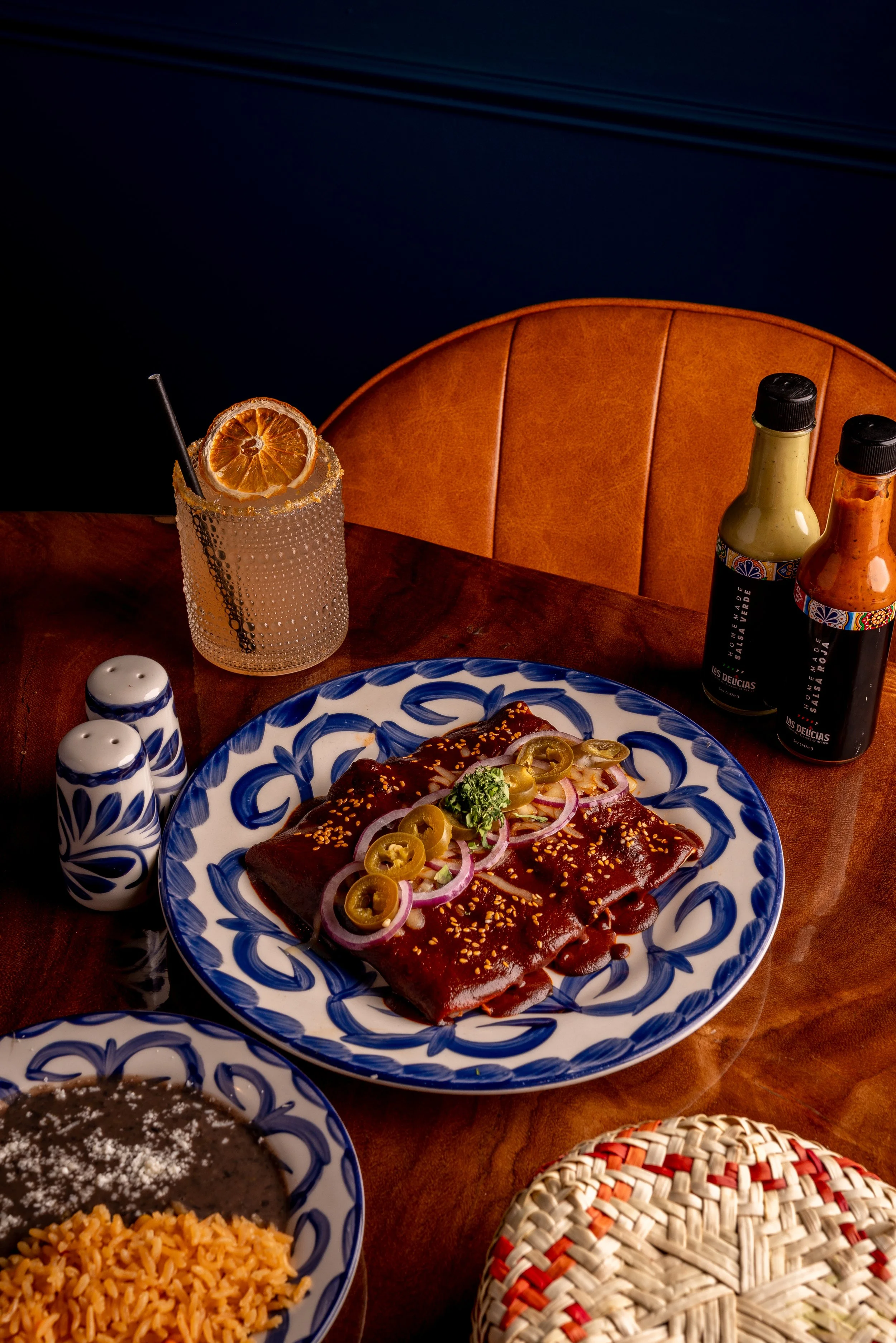 Mexican meal with rice, chocolate cake, and grilled meat with sauce and sliced jalapeños on a decorative blue and white plate, with condiments and a glass of orange drink garnished with a dried orange slice and a straw. There is a woven colorful hat and salt and pepper shakers on a wooden table.