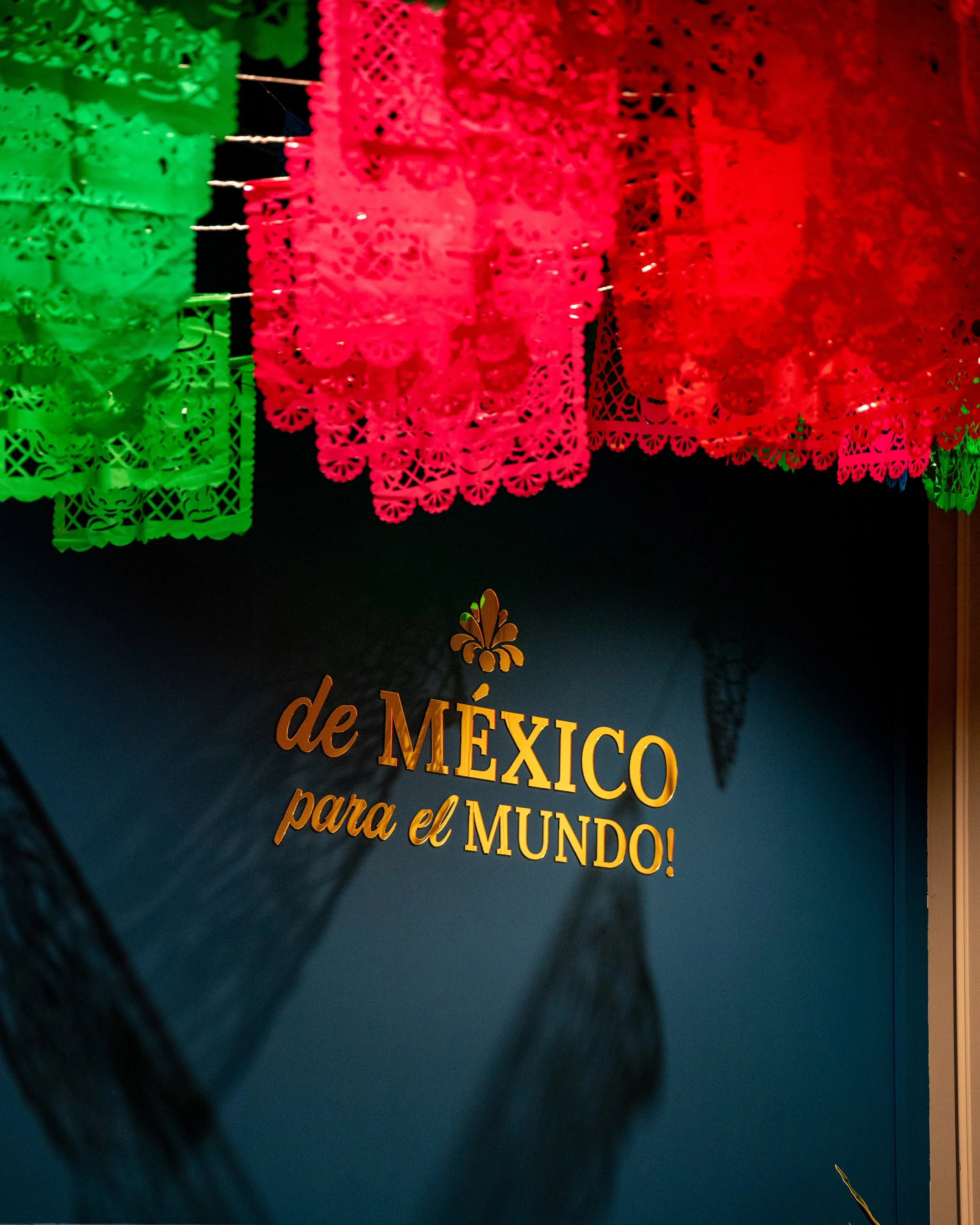 Colorful papel picado banners in green, red, and pink with Spanish text that says "de MÉXICO para el MUNDO!" against a dark background.
