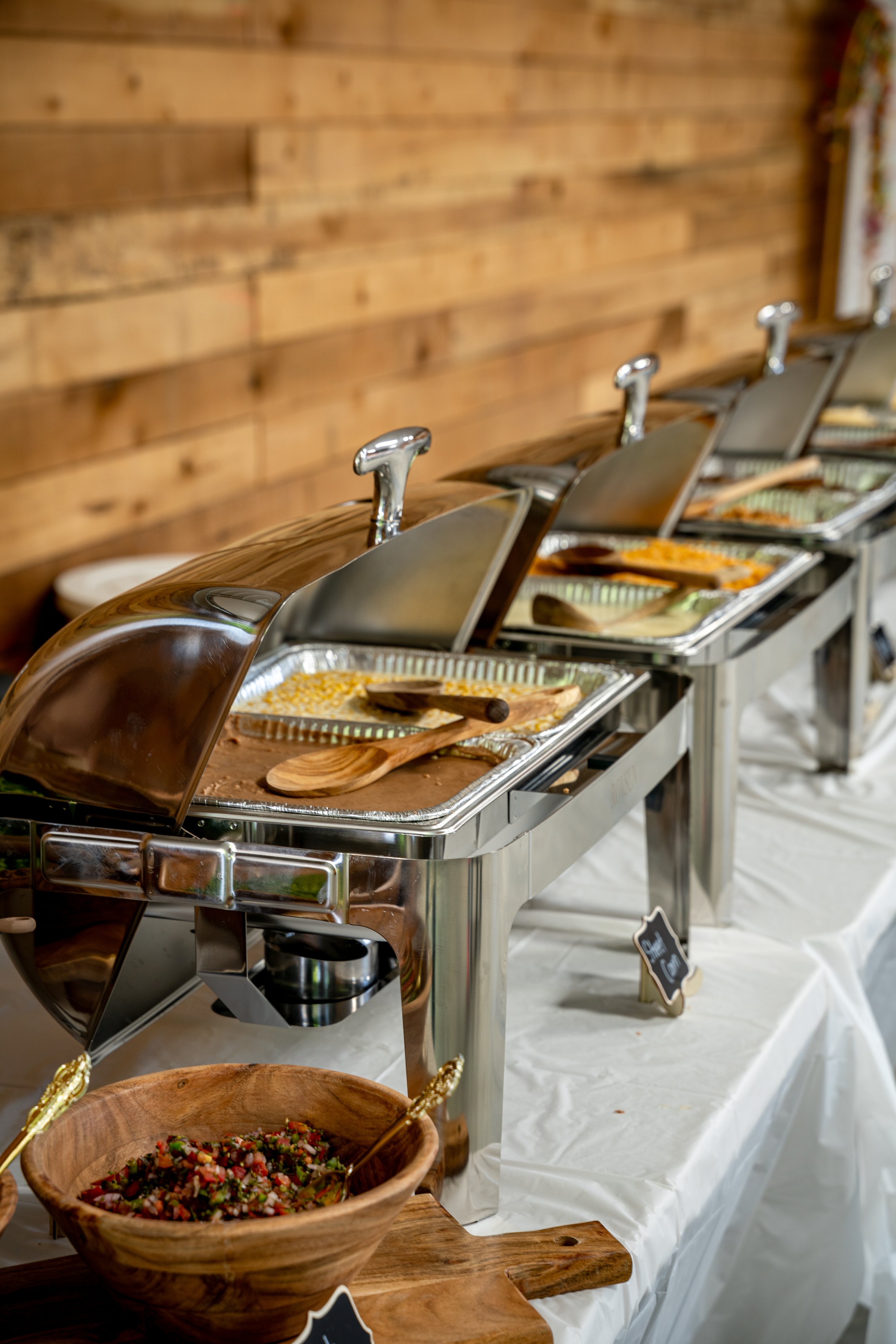 Buffet station with chafing dishes containing various hot foods, bowls with salads and toppings, set against a wooden wall with a white tablecloth.