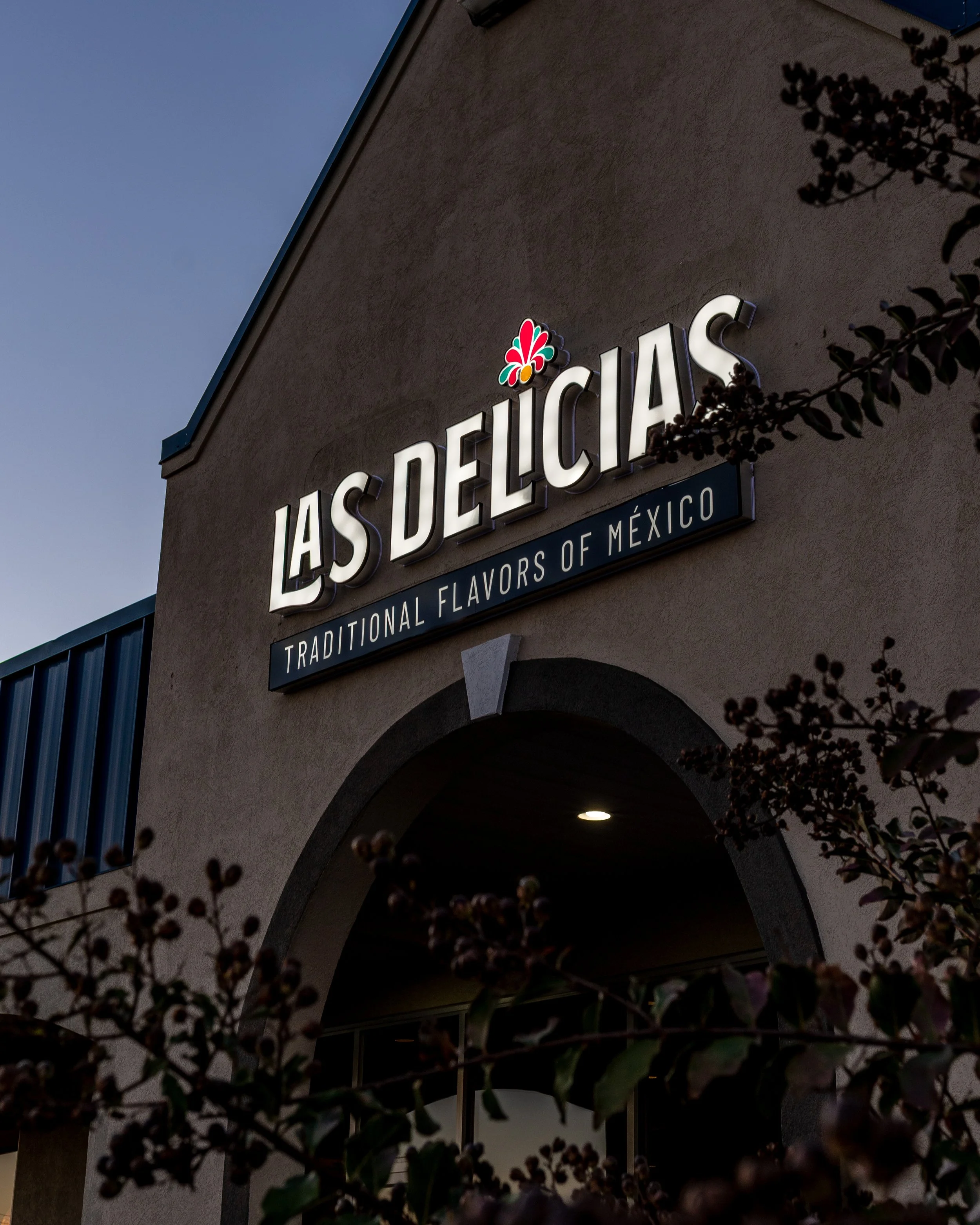 The exterior of a building at dusk with illuminated signage reading "Las Delicias" and a logo above the name, partially obscured by plant foliage in the foreground.