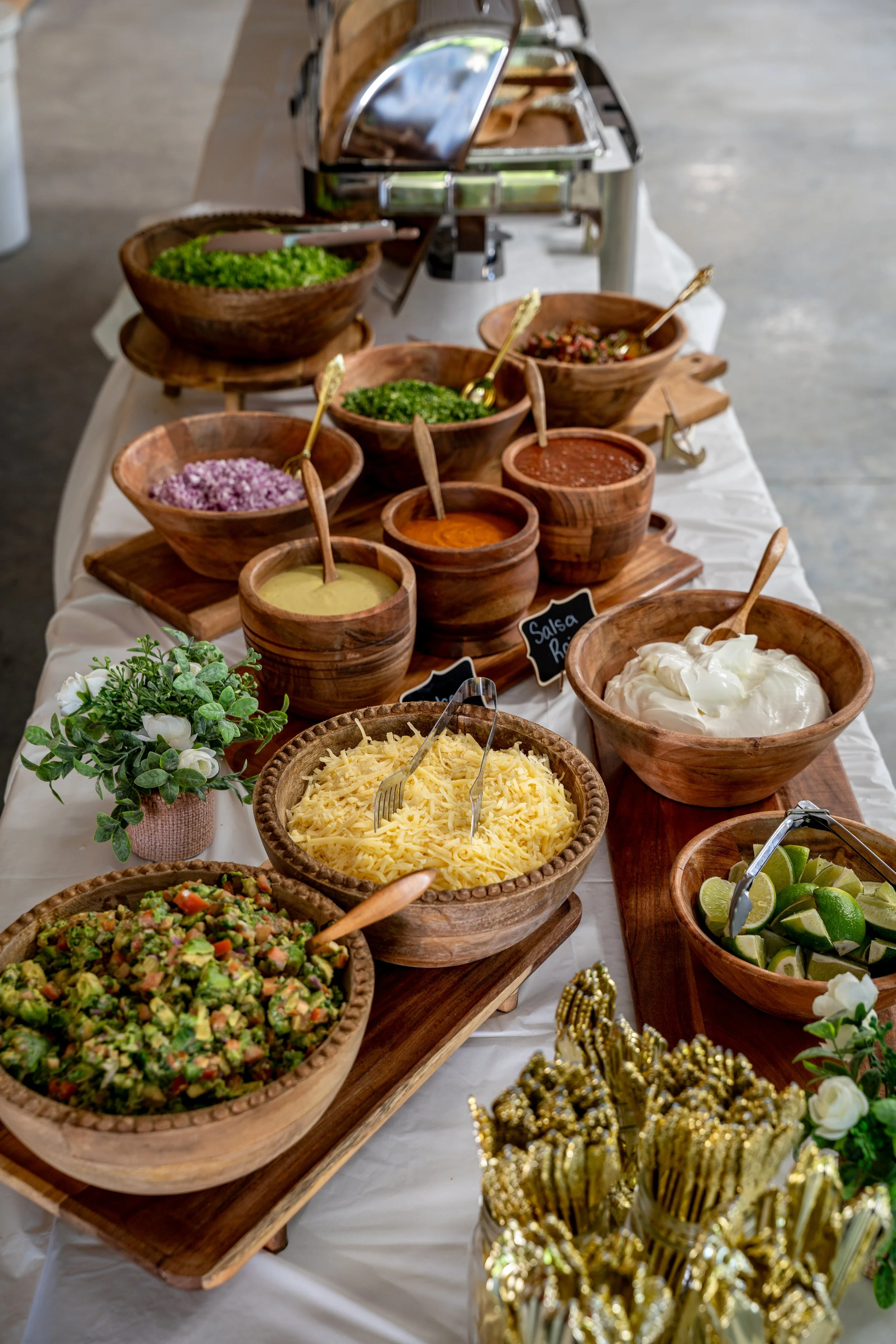 A buffet table with wooden bowls filled with various Mexican condiments like shredded cheese, guacamole, salsa, chopped vegetables, and lime wedges, garnished with white flowers and gold decorative accents.