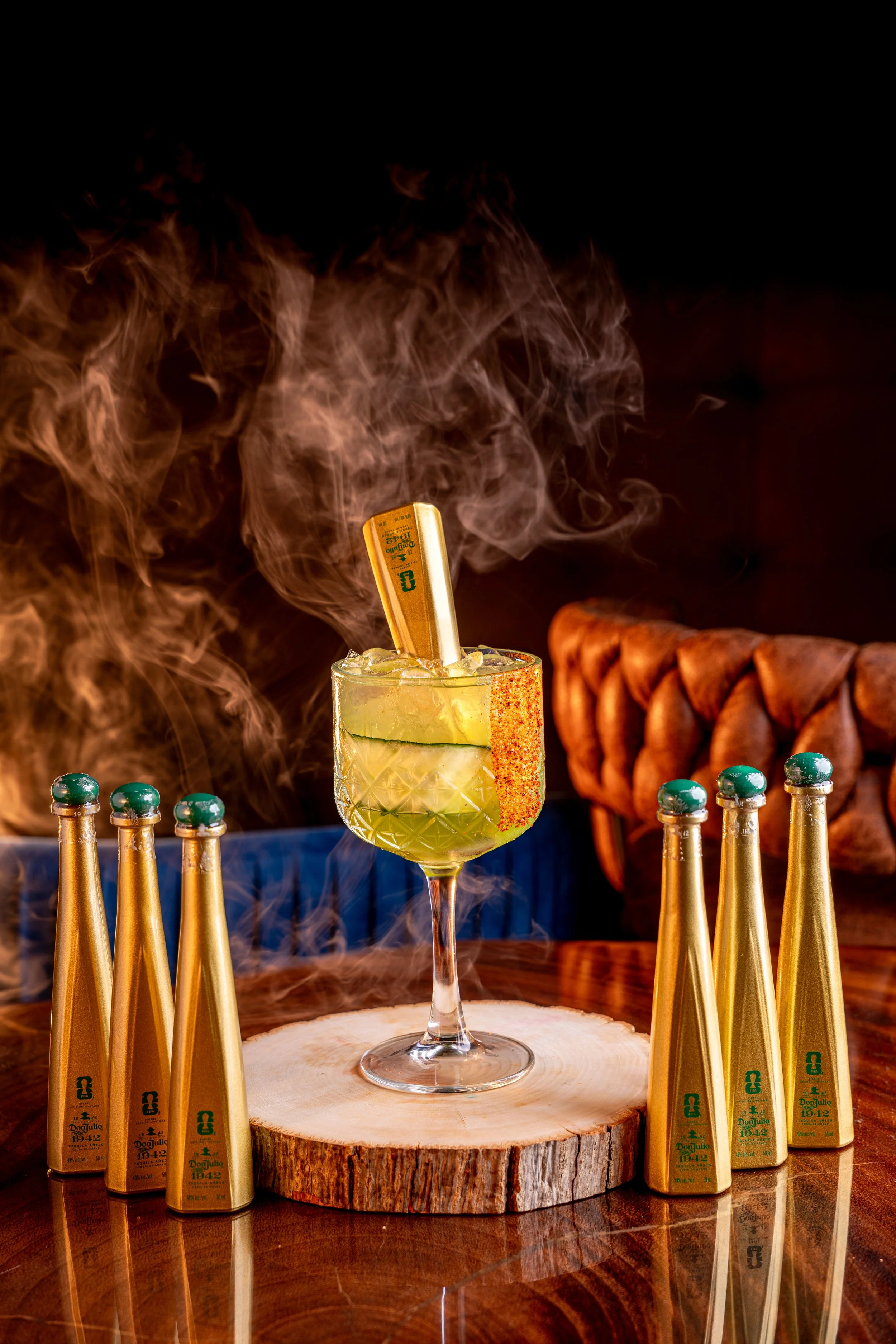A festive scene with a classic cocktail garnished with a cucumber slice, a spice-rimmed glass, surrounded by multiple bottles of Don Julio tequila, a carved wooden serving board, and a blurred background with warm lighting and a leather chair.
