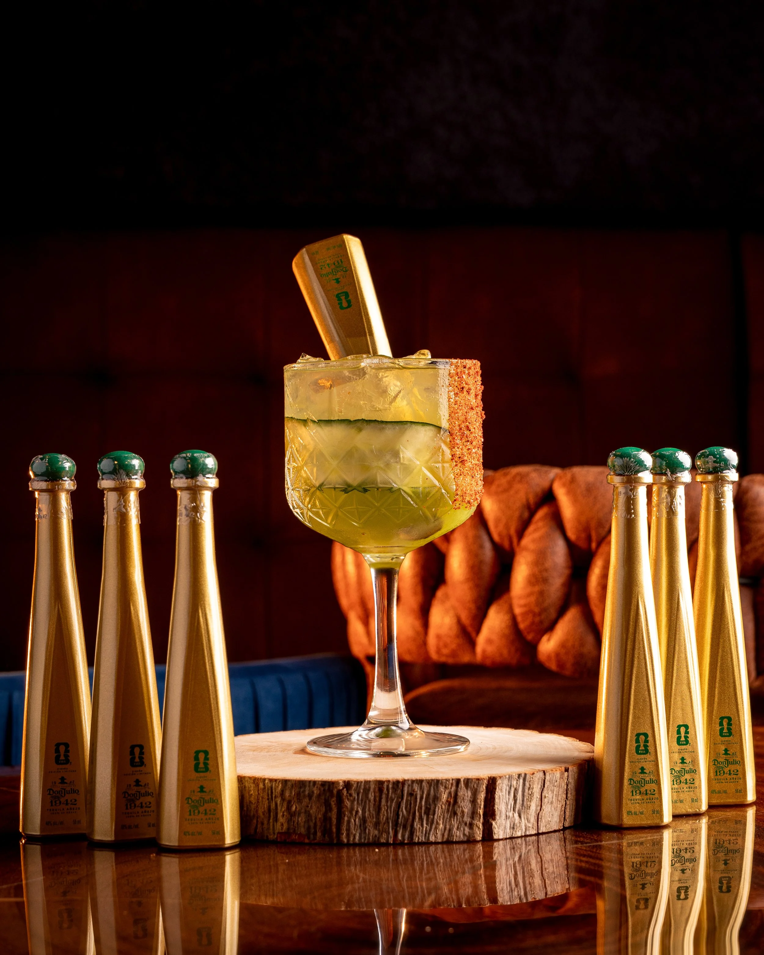 A yellow-green cocktail with ice and cucumber slices in a decorative glass, garnished with a small container of Tabasco sauce, surrounded by several bottles of Don Julio 1942 tequila on a wooden surface.