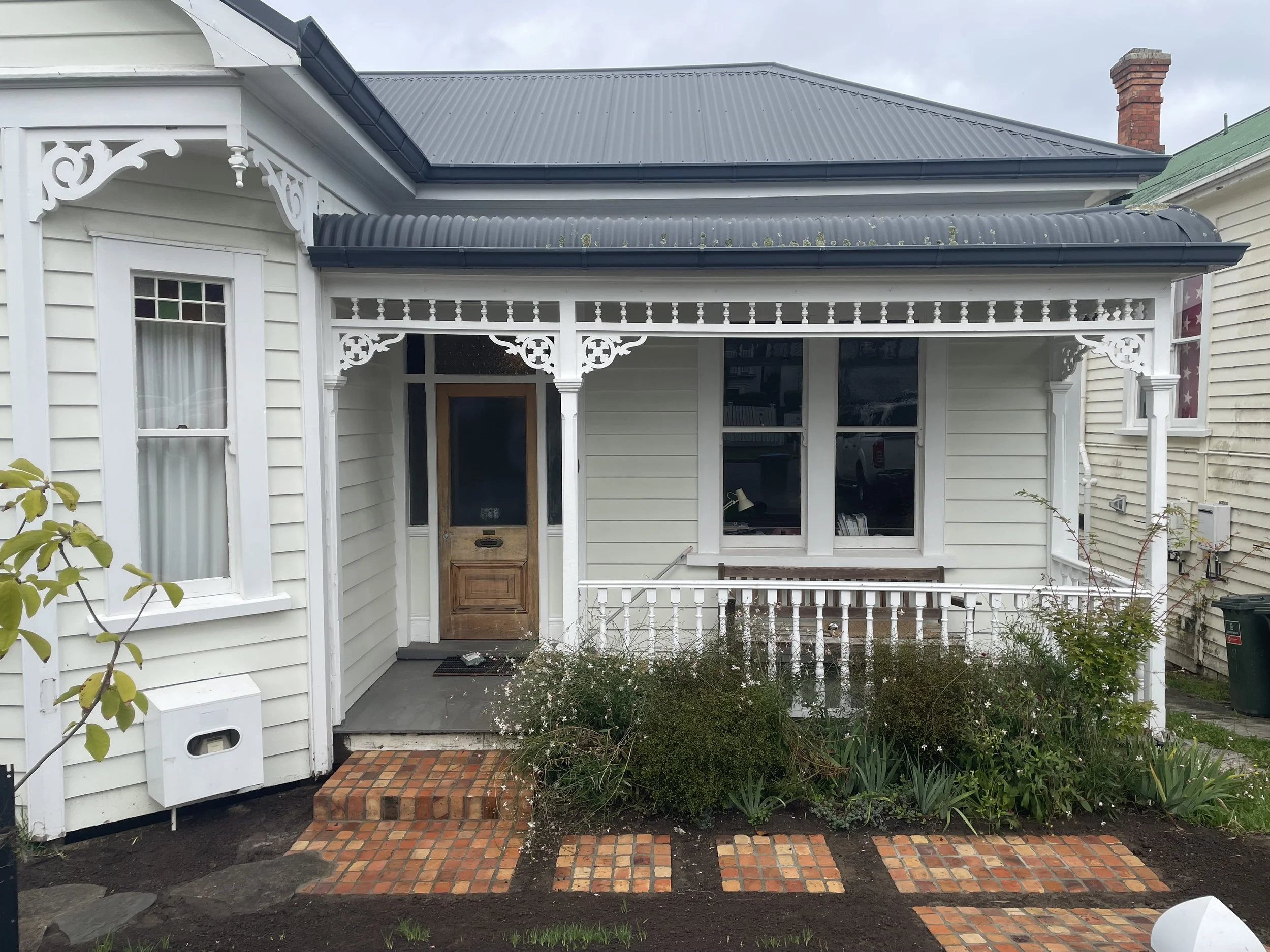 Front porch of a white Victorian house with decorative trim, a wooden front door, brick steps, a garden with plants, and a brick walkway.