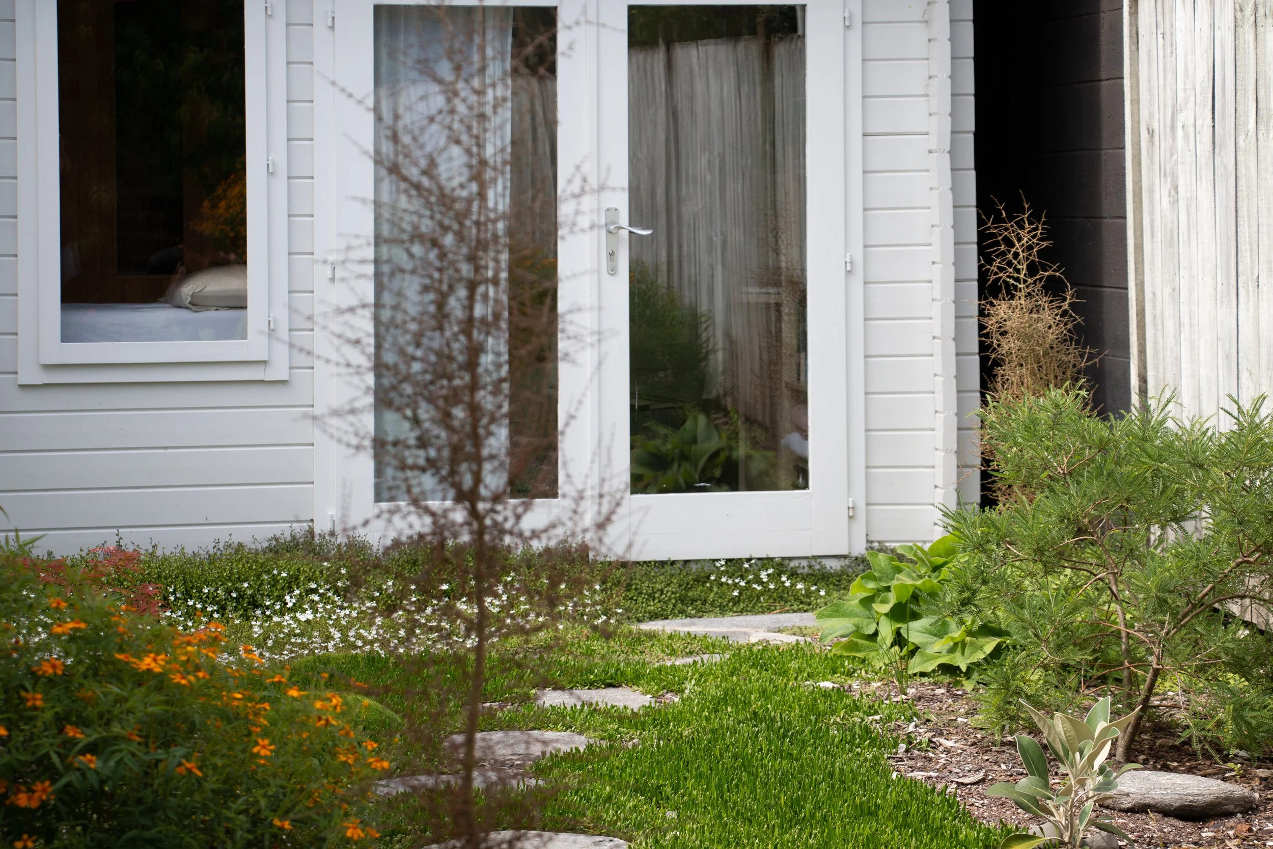 View of a house garden with a white door and window, surrounded by green plants, flowers, and small stepping stones.