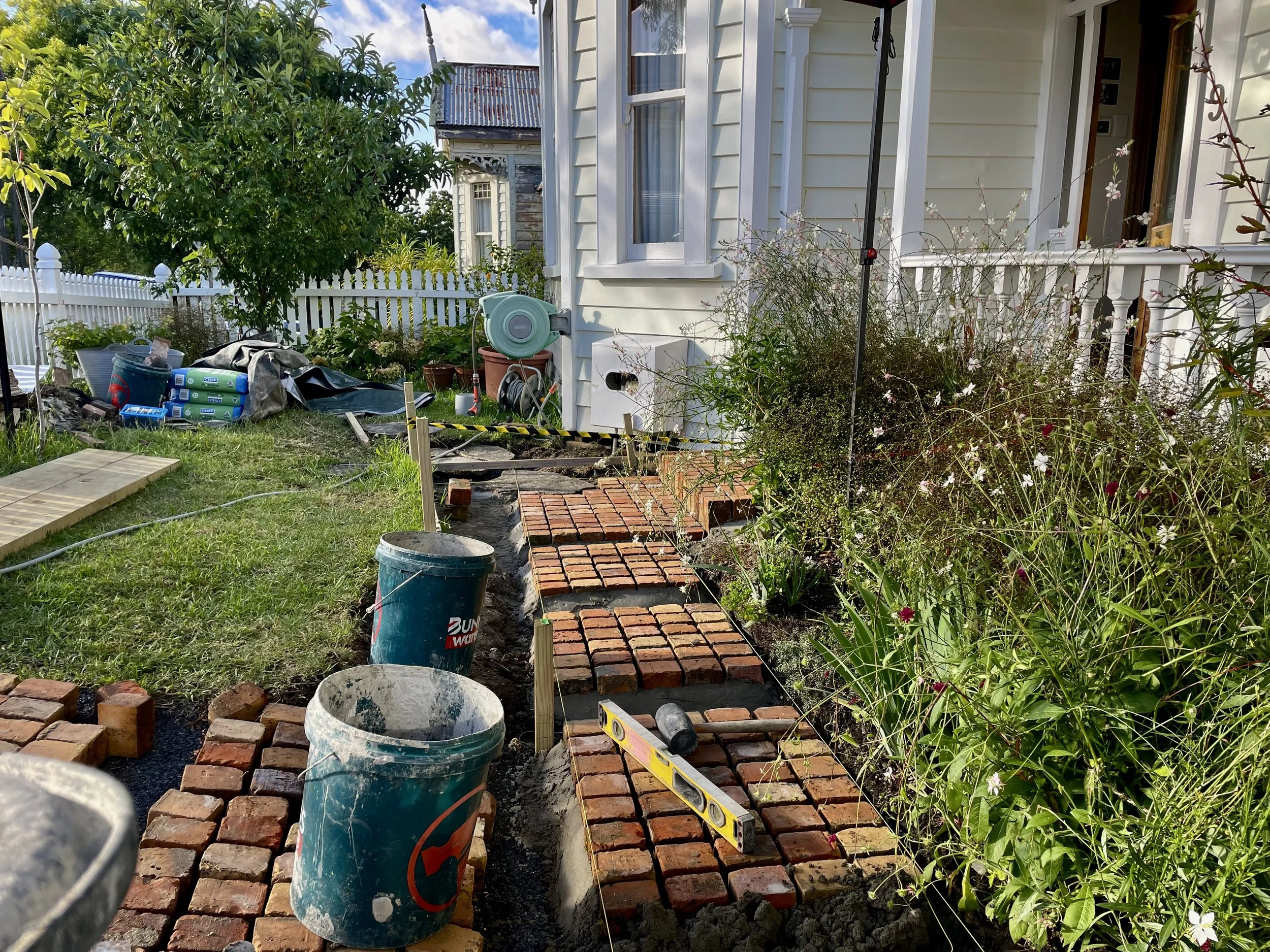 Construction site of a brick pathway in a garden next to a white house, with construction tools, buckets, and plants.
