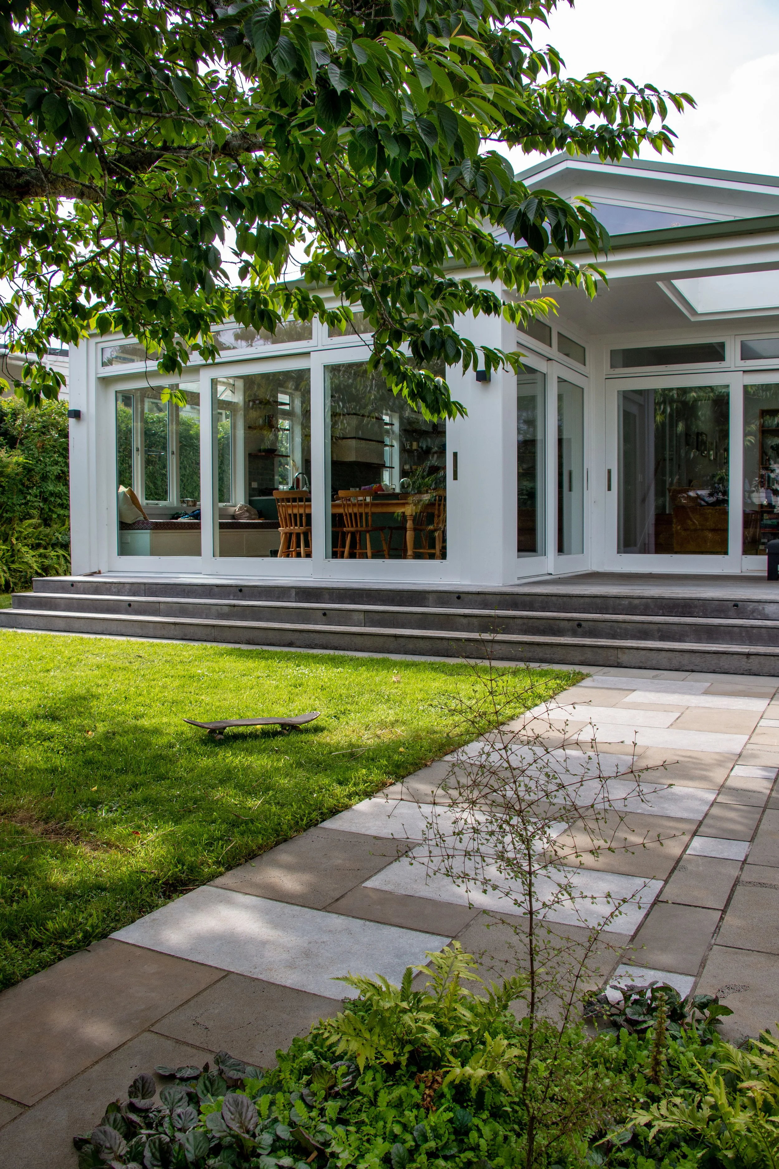 A modern house with large glass windows and a deck, surrounded by a lush green garden with a skateboard on the grass and a stone pathway.