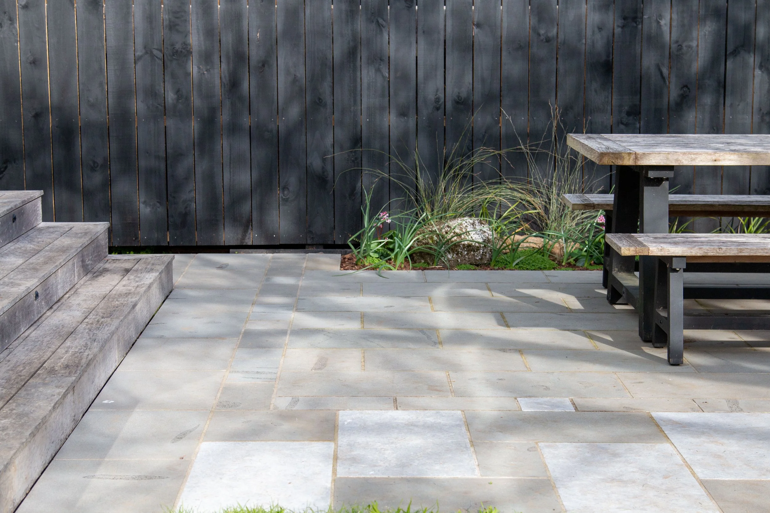 A patio with stone pavers, wooden stairs on the left, a dark wooden fence in the background, and a wooden picnic table with benches on the right. There is a small garden bed with plants and rocks near the fence.