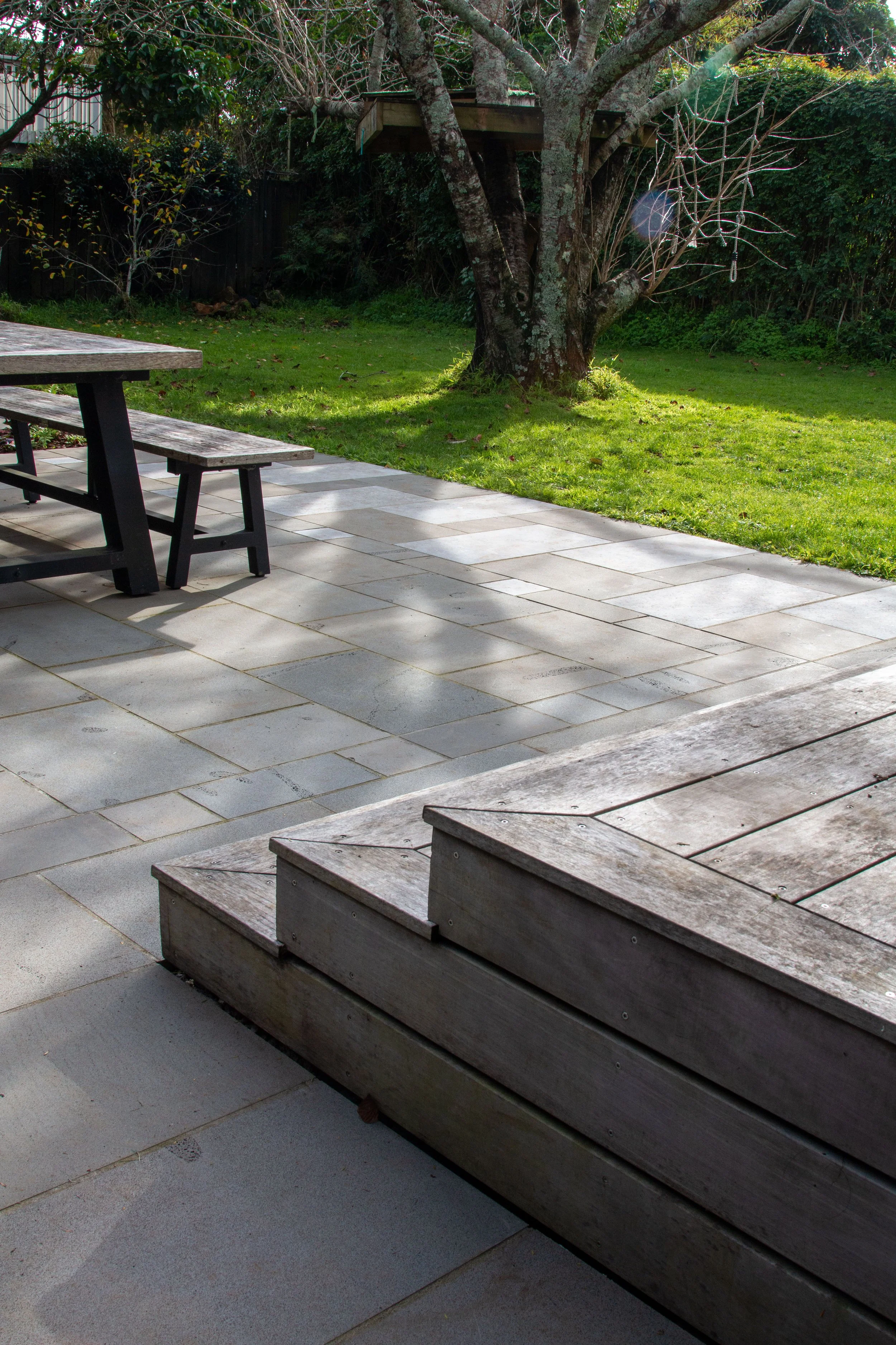 A backyard patio with stone flooring, a wooden deck with steps, a picnic table, and a large tree with a treehouse