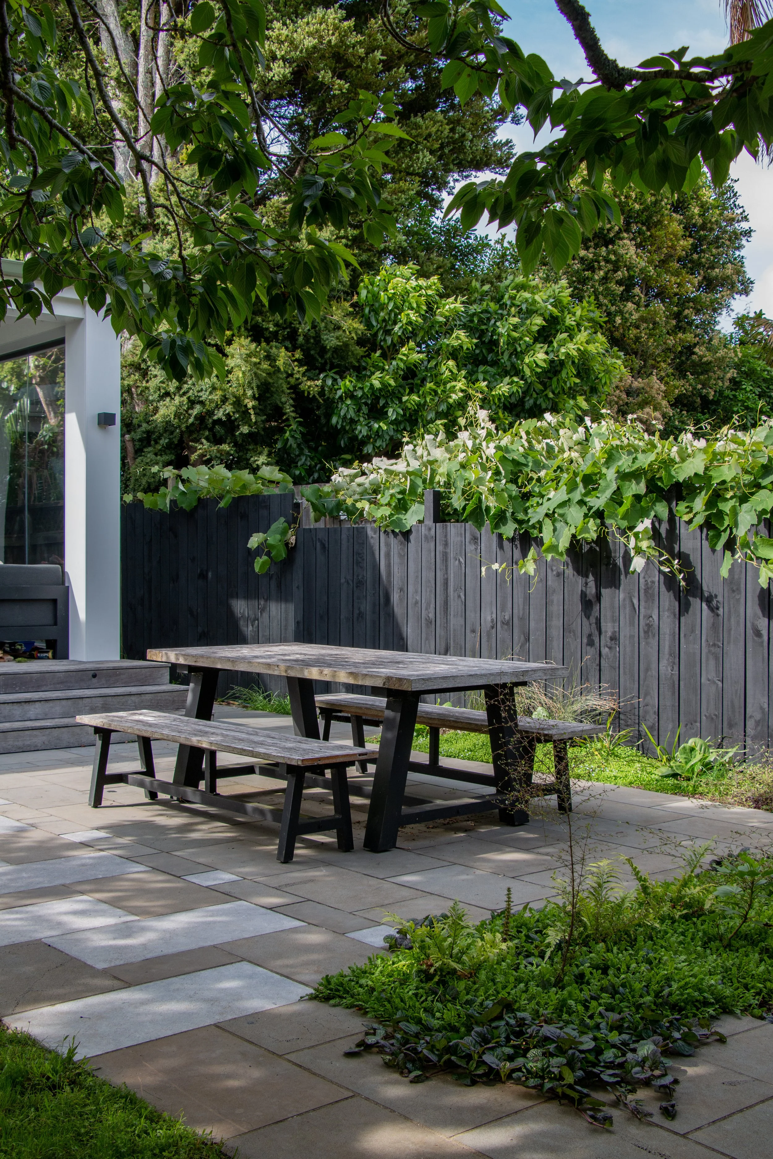Outdoor patio with a wooden table, benches, and a small garden area surrounded by lush trees and plants in a backyard.