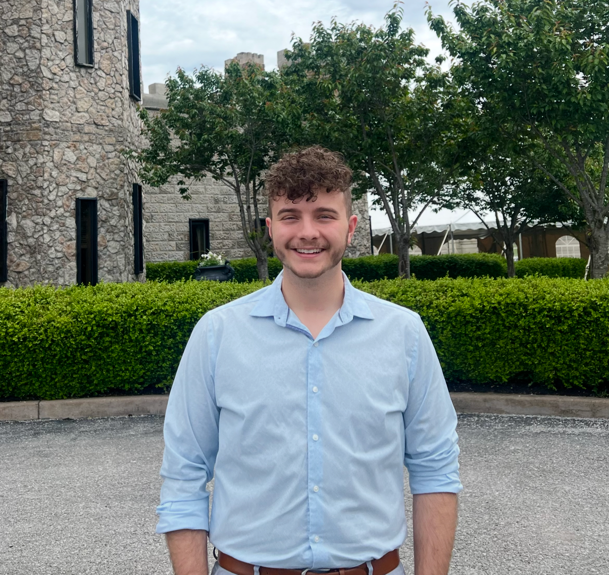 A young man with curly brown hair and a beard smiling outdoors, wearing a light blue button-down shirt with the sleeves rolled up, standing in front of a row of green bushes and trees, with a stone building and a tent in the background.