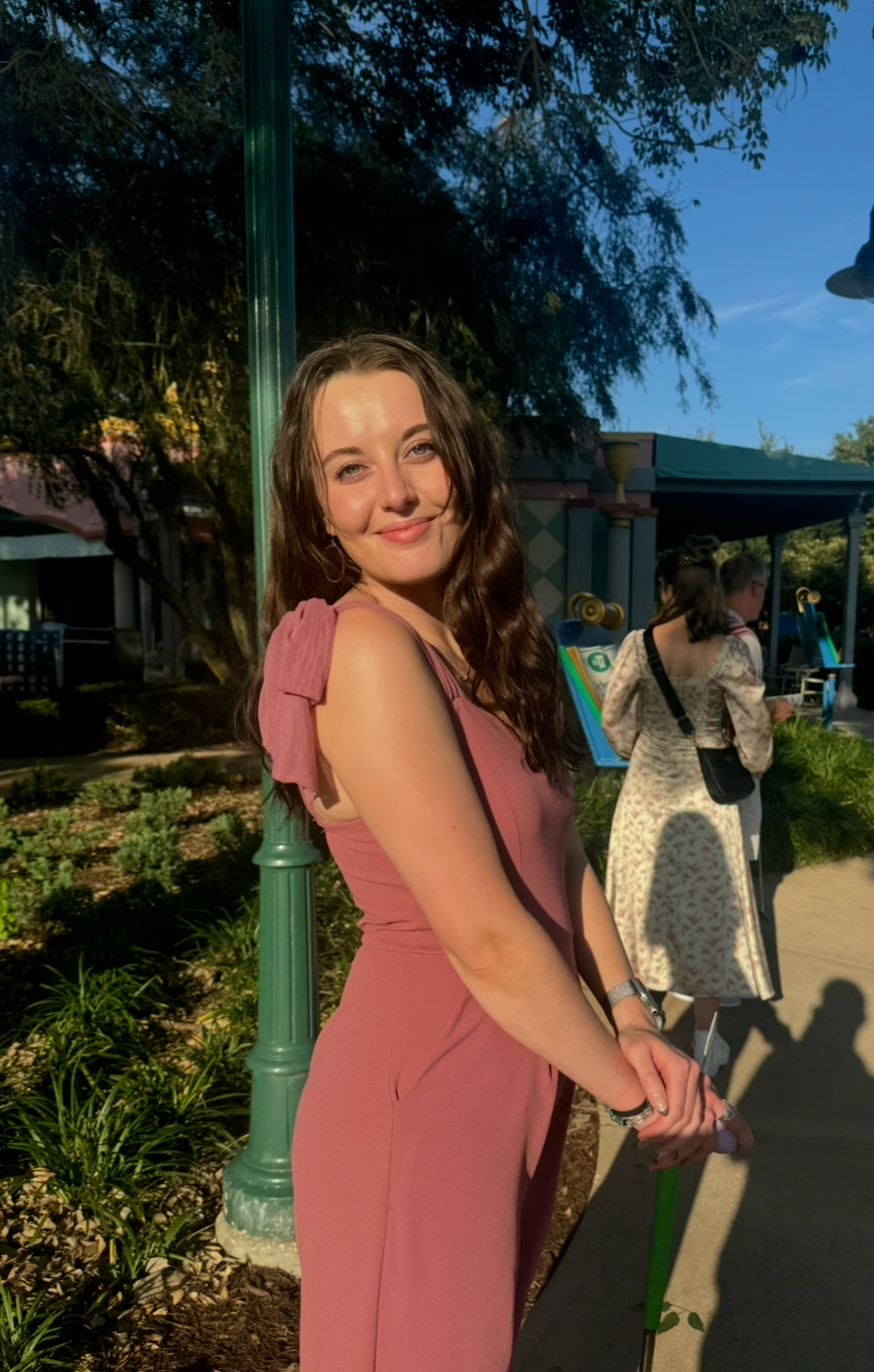 A young woman with long, wavy brown hair, wearing a mauve sleeveless dress with ruffled shoulder details, is smiling at the camera. She is outdoors in a park-like setting with other people in the background, and the sun is shining, casting shadows.