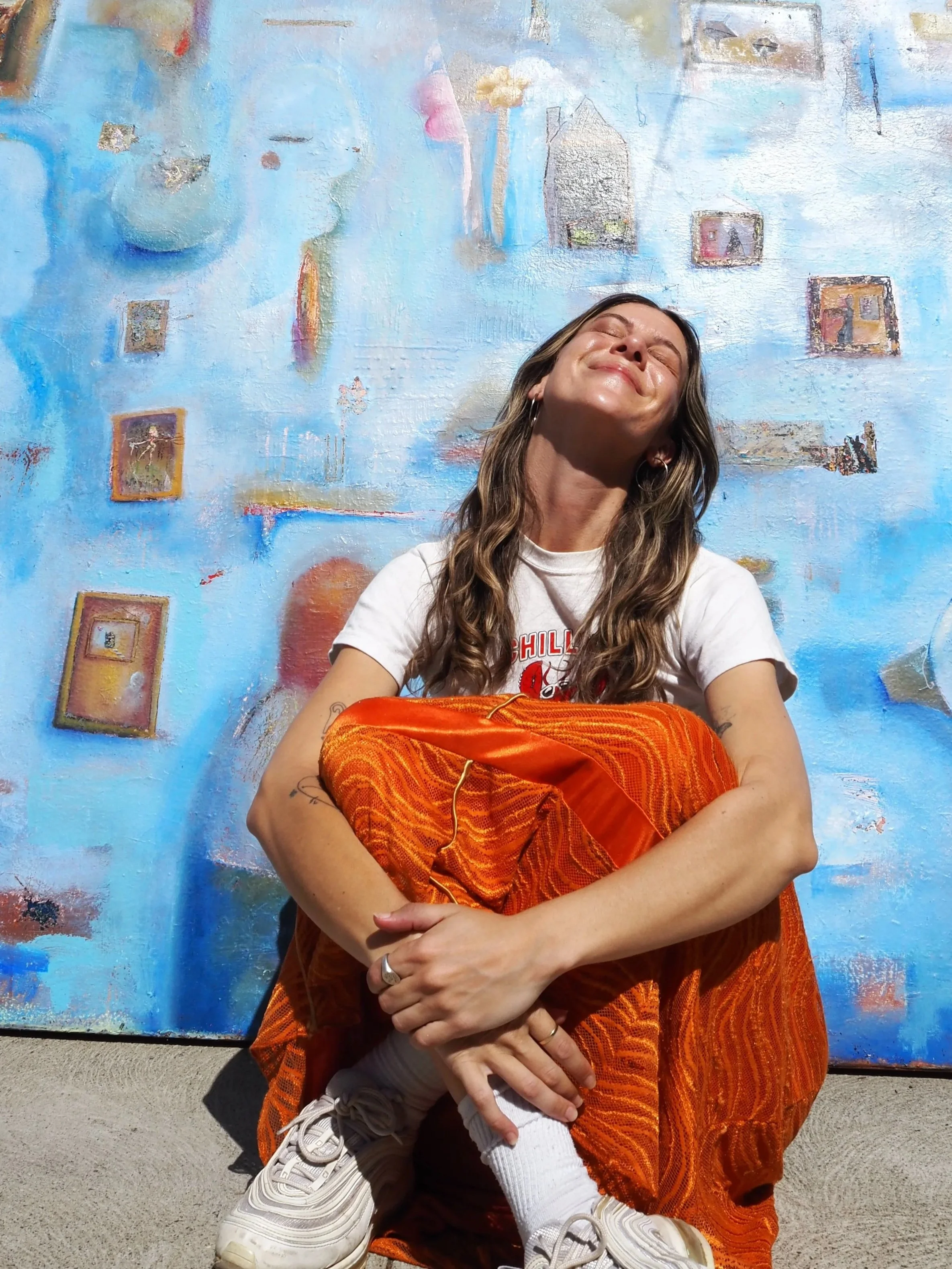 A woman with long wavy hair, wearing a white T-shirt, orange patterned pants, and sneakers, sitting on the ground against a colorful abstract painted wall, smiling with eyes closed.