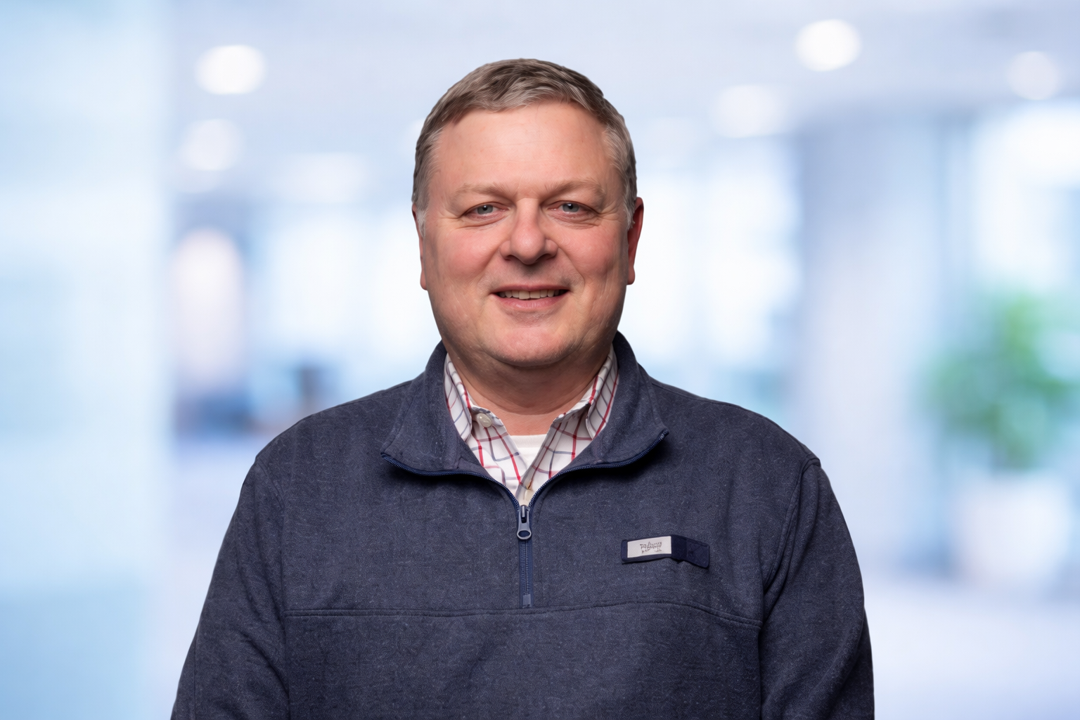 Al Bell is the owner of Belltower Church Consulting. A smiling man with short light brown hair wearing a navy blue quarter-zip fleece and a white and red plaid shirt standing in an office with a blurred background.