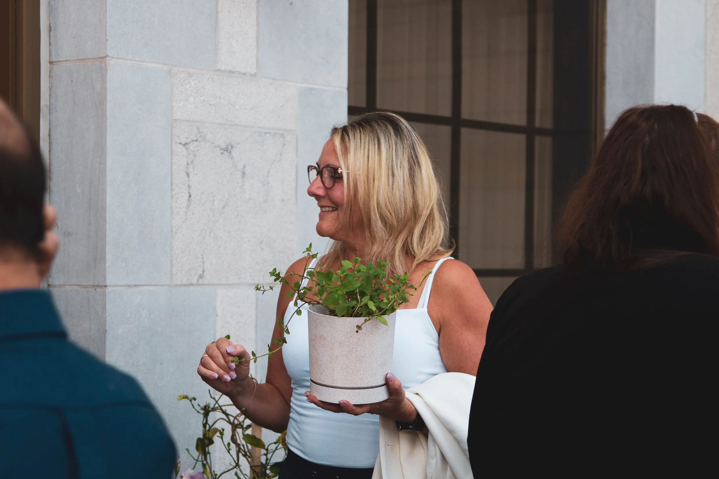 A woman with blonde hair and glasses holding a potted plant with green leaves, smiling and wearing a white top and a beige jacket, talking to a group of people outdoors near a light-colored building.