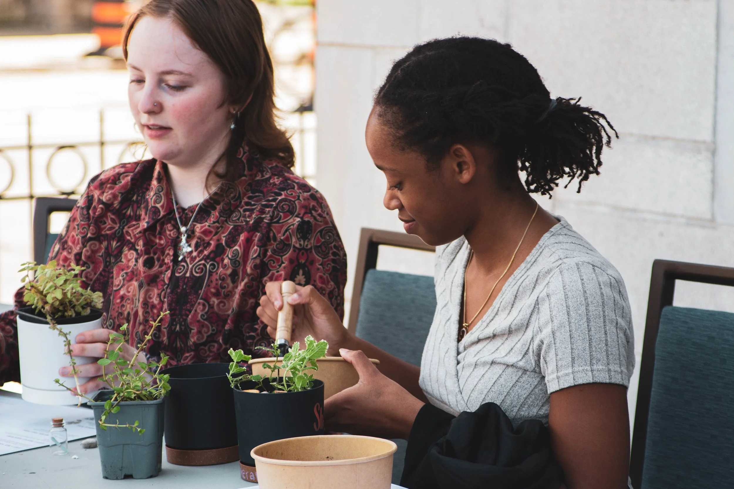 Two women are sitting at a table planting and caring for small potted plants, engaging in gardening activities.