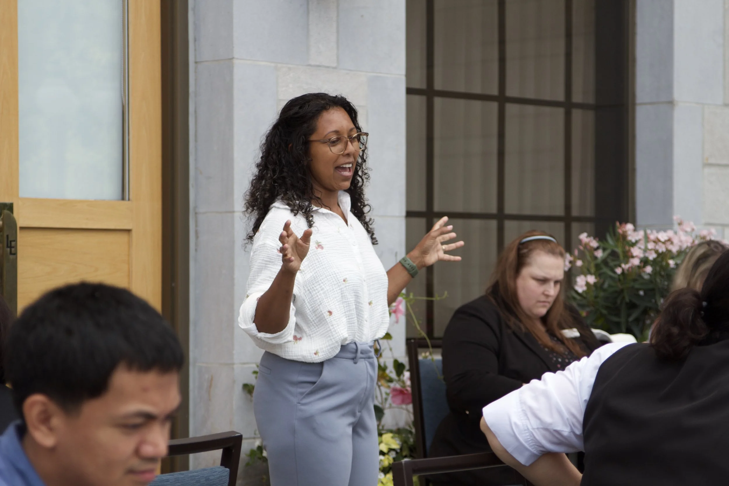 A woman with glasses and curly hair, wearing a white blouse, speaking to a group of people seated outdoors near a building with large windows and flowers.