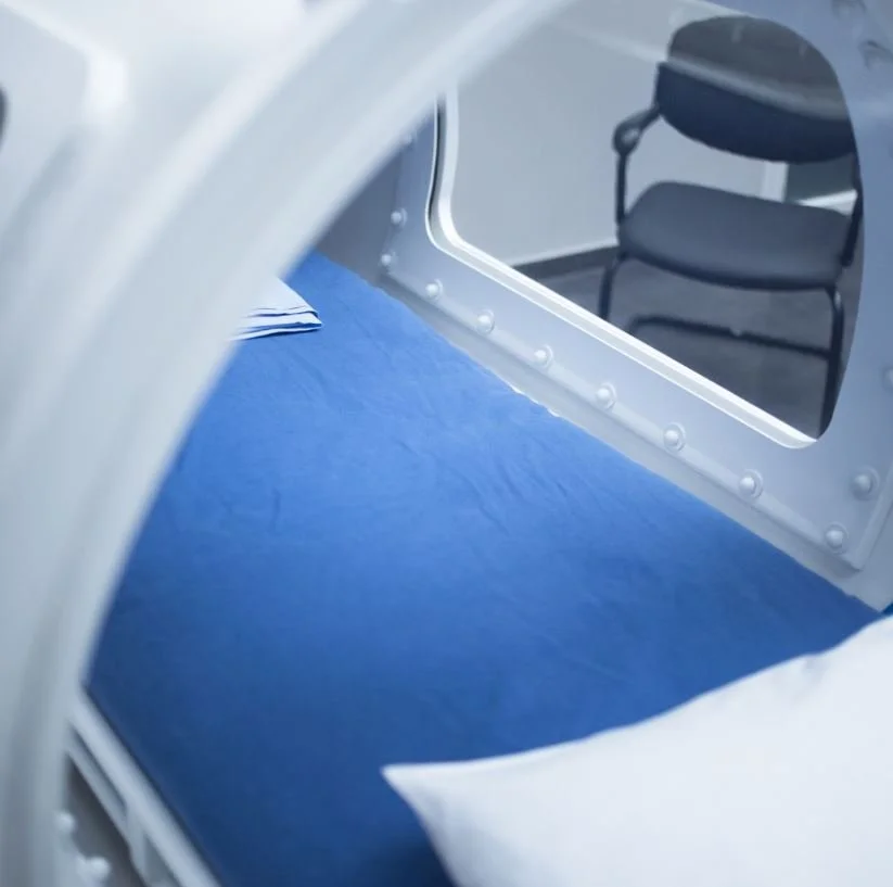 View inside a spacecraft or capsule showing a blue mattress, a white pillow, and a chair with a cushion in the background.