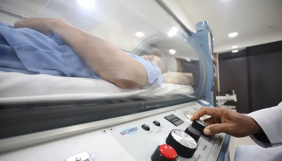 A person lying inside a medical imaging machine, possibly an MRI or CT scanner. A technician operates the machine controls.