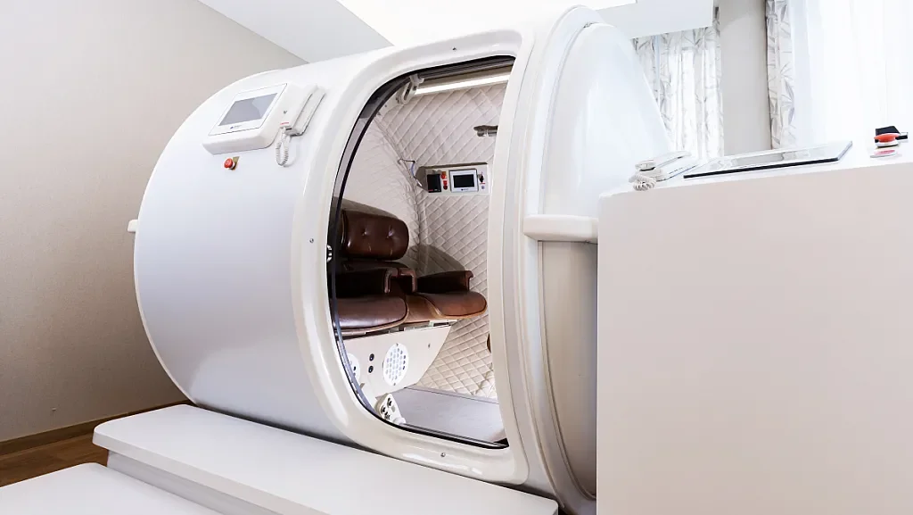 A modern hyperbaric oxygen chamber with a brown leather chair inside, set in a white room with curtains.