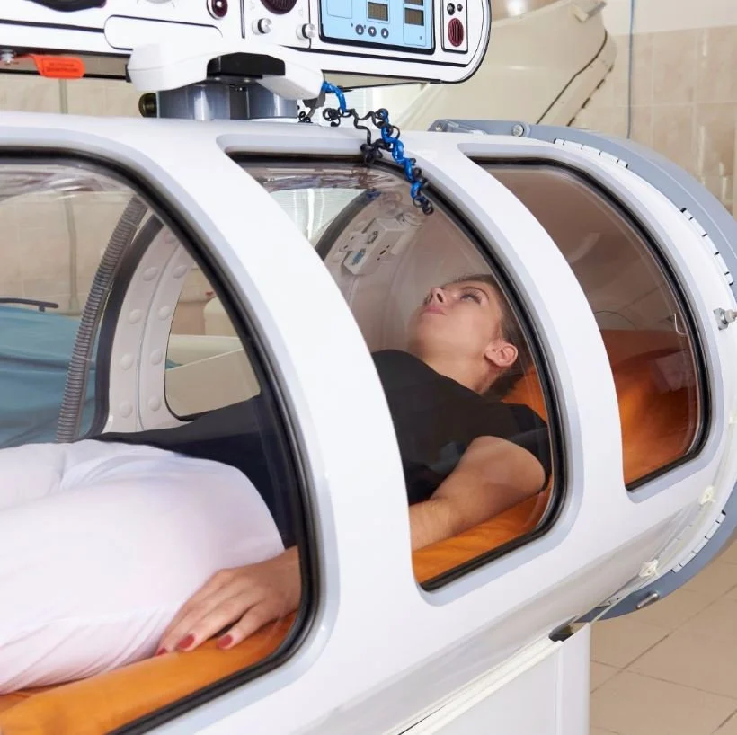 Woman inside a hyperbaric oxygen therapy chamber lying on a bed, looking up through the clear chamber window.