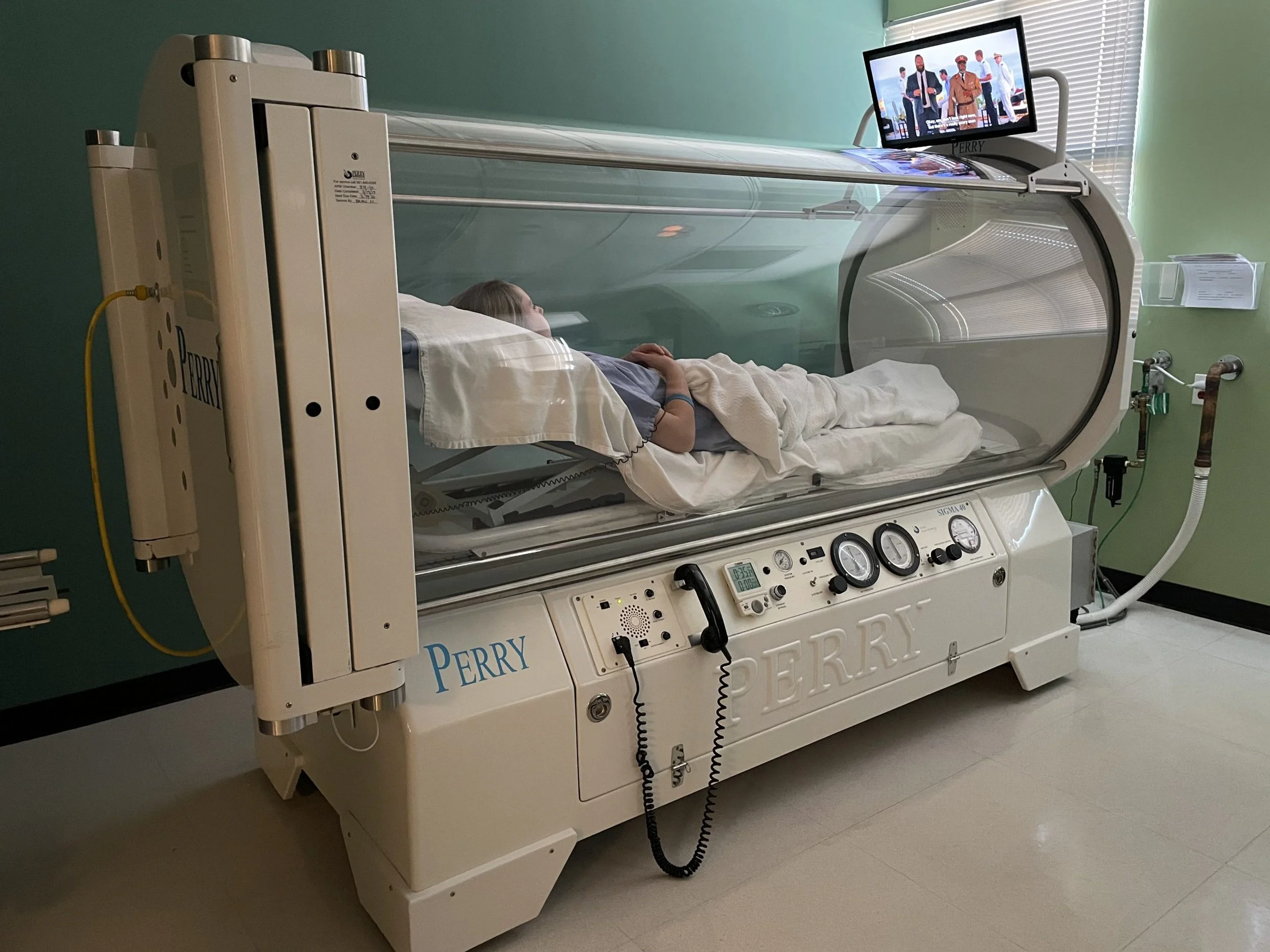 Child lying inside a hyperbaric oxygen therapy chamber, connected to the machine with monitoring controls on the front, in a medical room with a television on top of the chamber displaying a group of people.