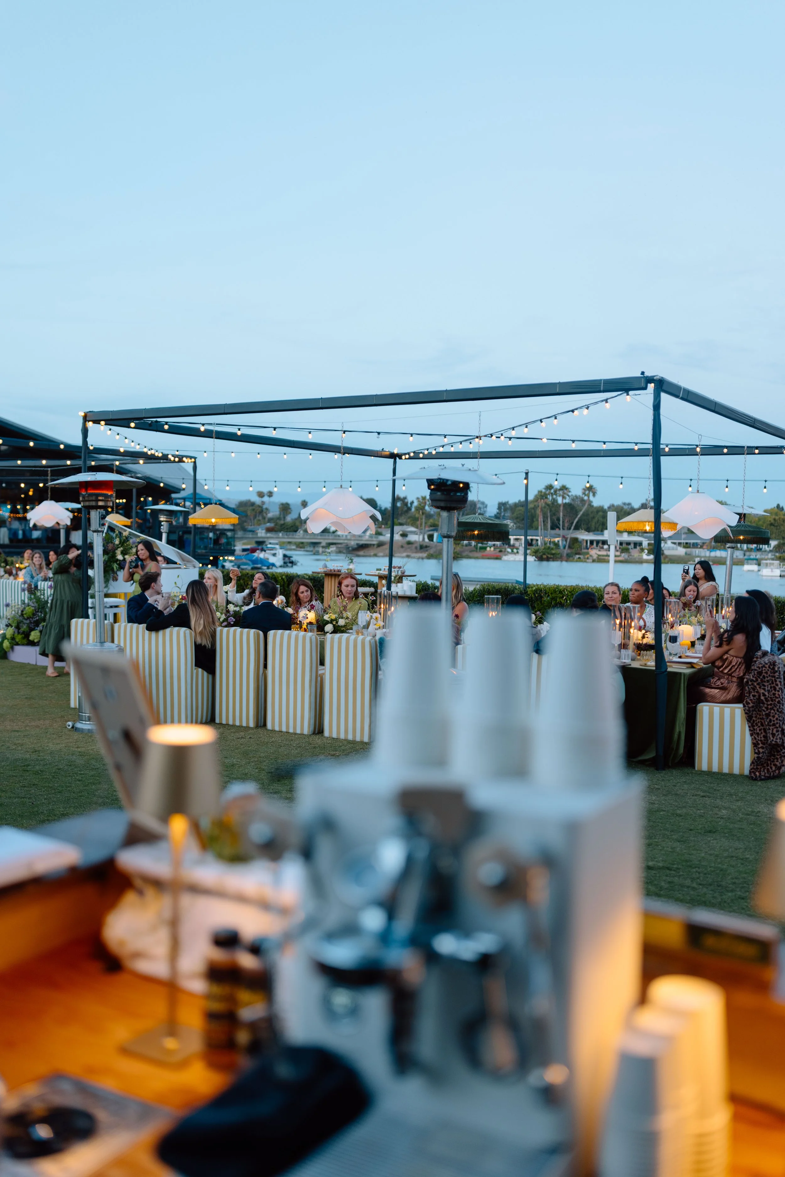 An outdoor gathering with people seated at tables, string lights overhead, and a scenic water view in the background during the evening.