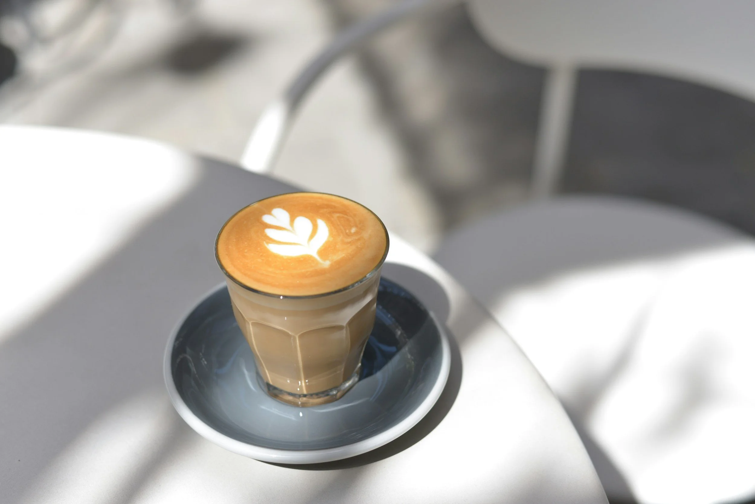 A glass of latte with latte art on top, placed on a saucer on a white table, with shadows and a blurred background.