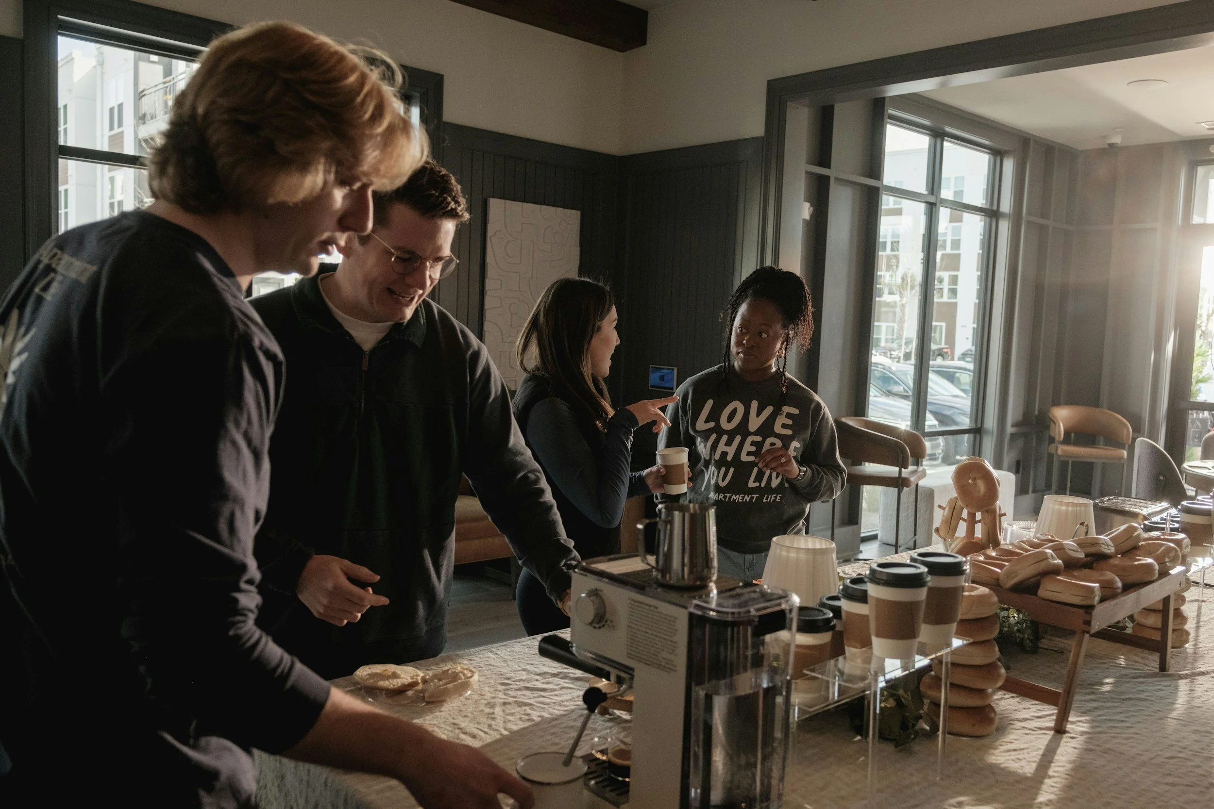 People gather around a breakfast buffet table with coffee cups, bread, and a juice machine in a sunlit room with large windows and modern decor.