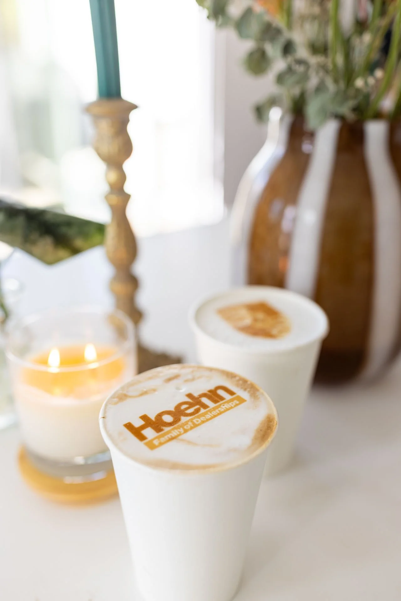 Three white cups of coffee with foam on top, one with the 'Harmon's Family of Dealerships' logo, a glass with a lit candle, a small plate, and two brown vases with green plants in the background.