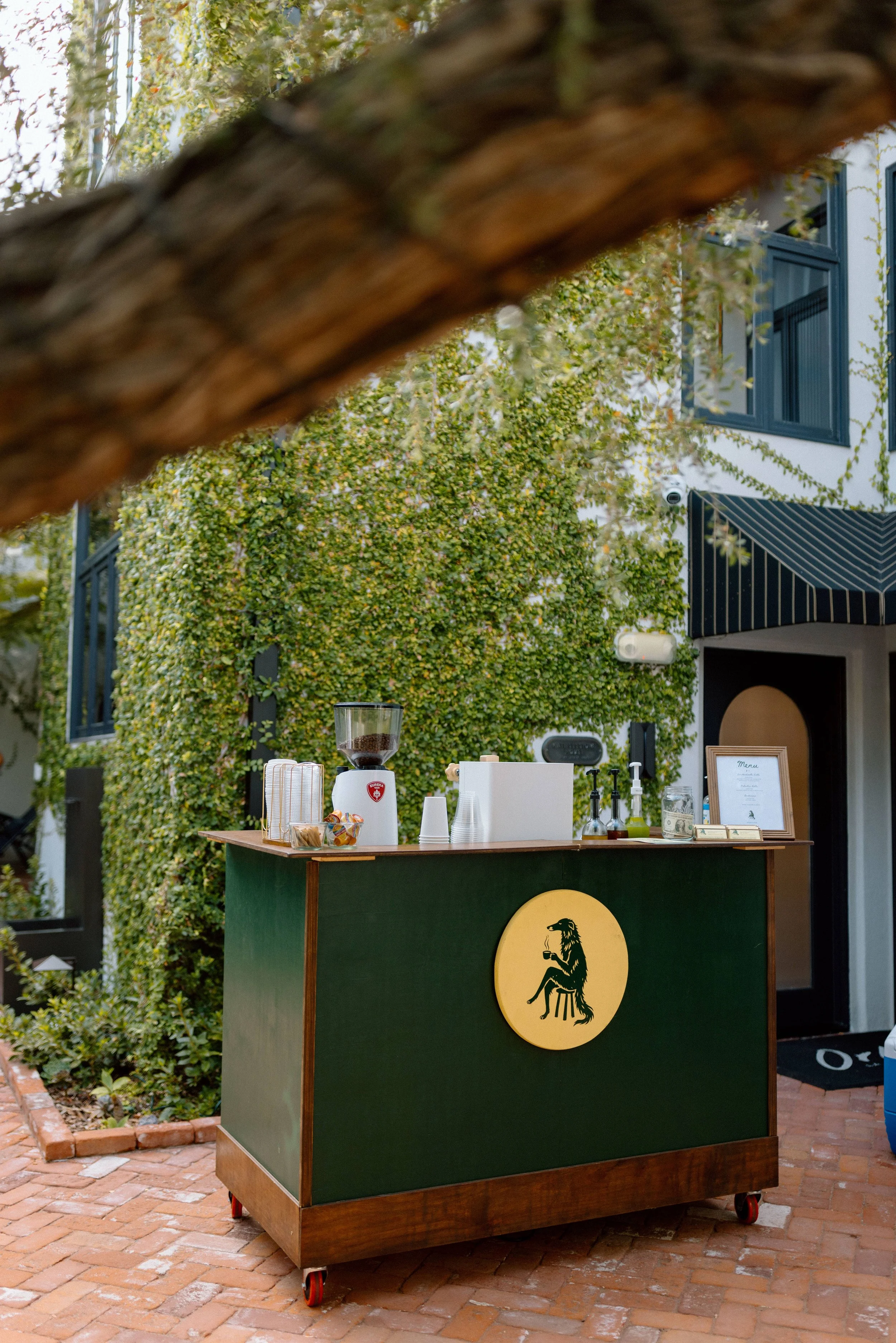 Outdoor bar or refreshment station with a green front featuring a yellow circular logo of a lion holding a glass, situated on red brick paving. The bar has various bottles, cups, and a coffee grinder on top, with a white framed menu and dollar bills visible. Background includes greenery and a white building with blue-framed windows and a striped awning.