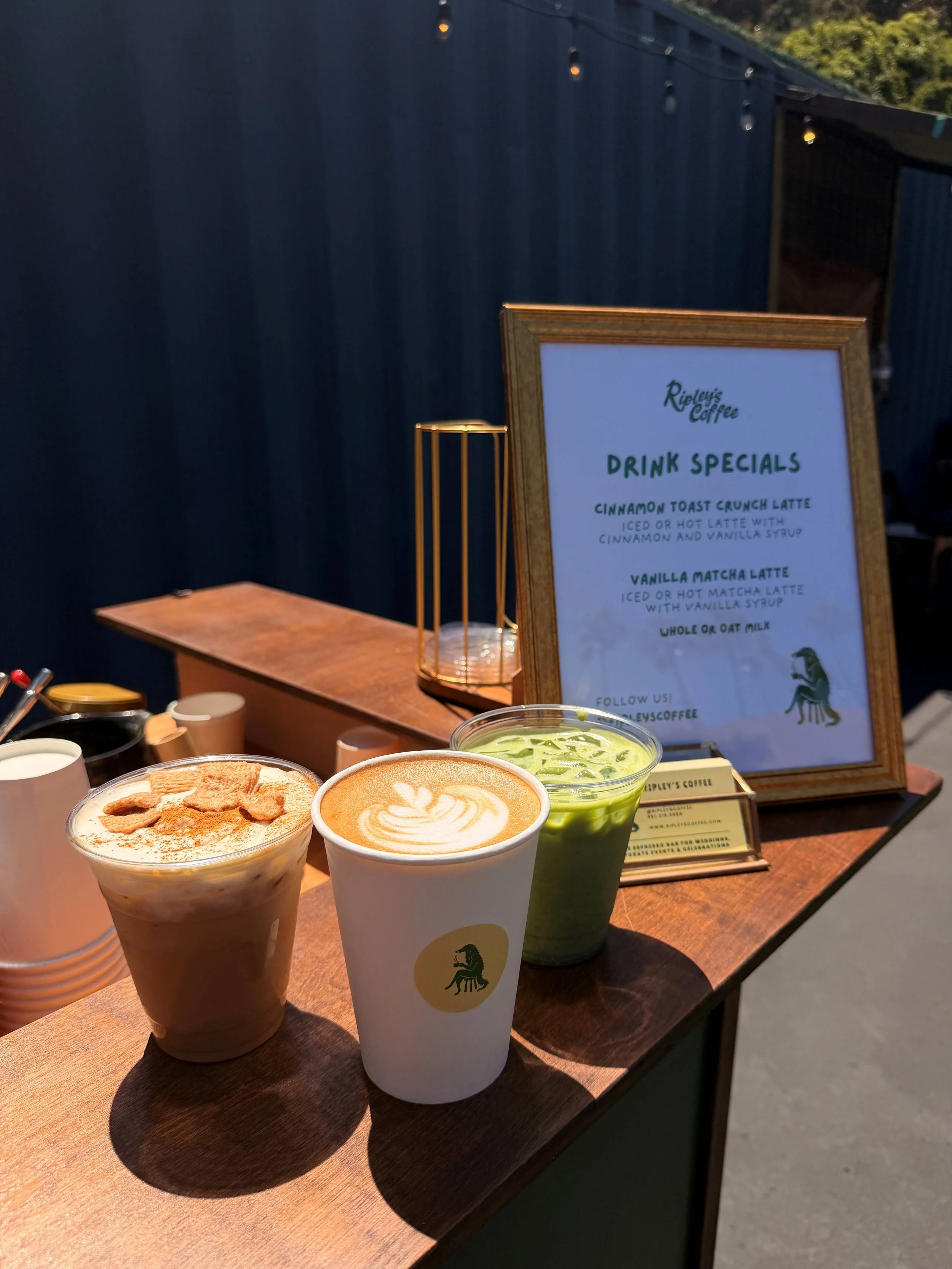 Three coffee drinks on a wooden table, with a sign in the background advertising drink specials, including cinnamon toast crunch latte, vanilla matcha latte, and whole or oat milk.