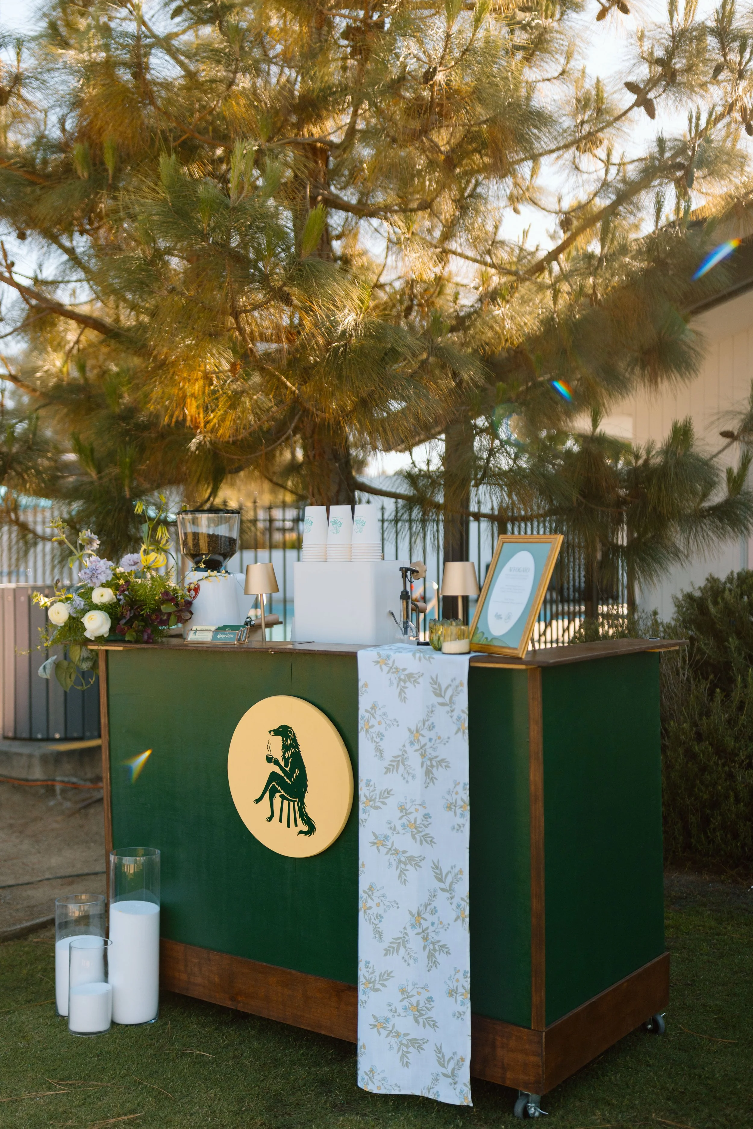 A green bar stand with a large yellow circle featuring a black dinosaur holding a coffee cup, flowers, candles, and framed menus or signs, set outdoors under a large tree.