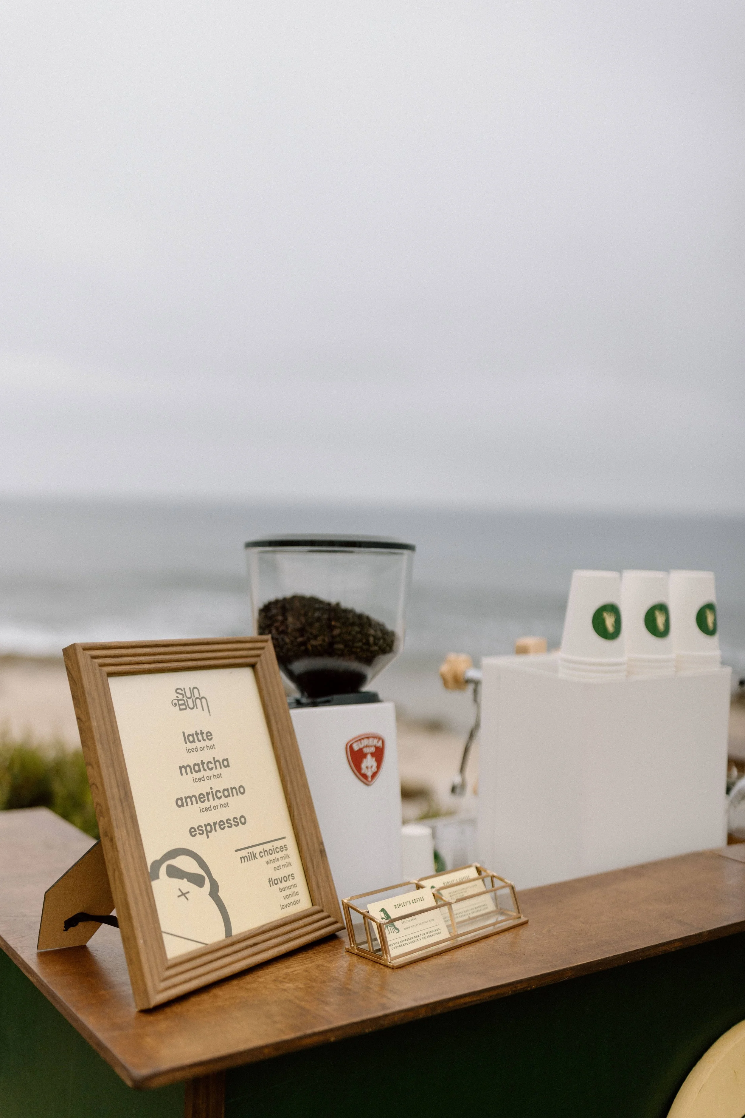 A close-up of a coffee stand with a wooden menu board listing different coffee drinks and a glass coffee grinder, with a white container holding disposable cups in the background.