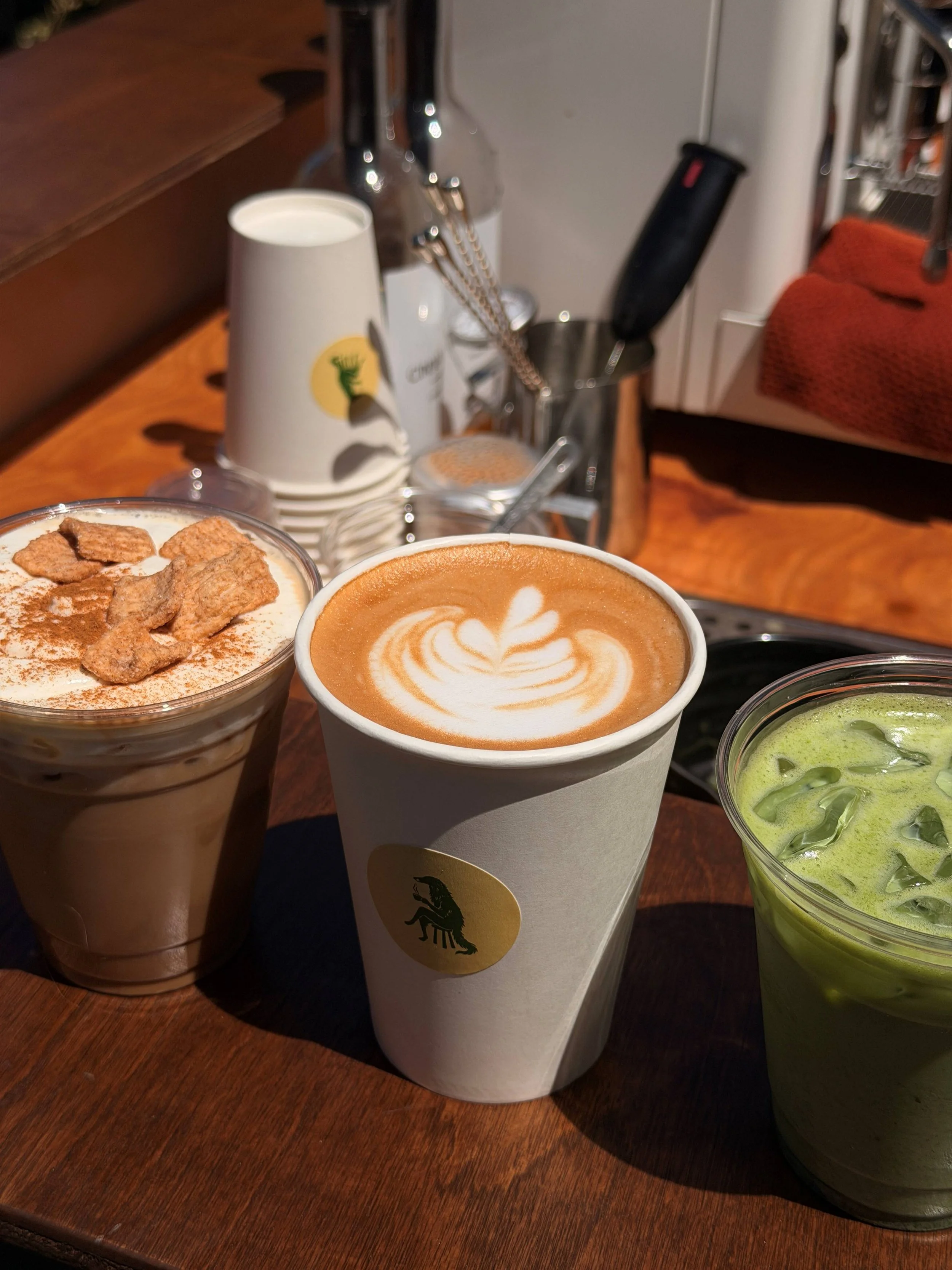 Close-up of various coffee drinks and a matcha latte on a wooden table, with one drink topped with whipped cream and cereal, another with latte art, and a third green matcha latte with ice.