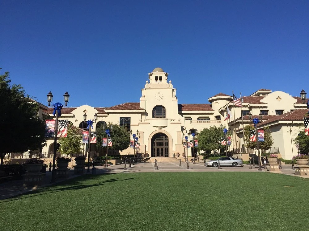 A large, white, historic-looking building with a clock tower, decorated with blue bows and American flags, with a well-maintained lawn and trees in the foreground.