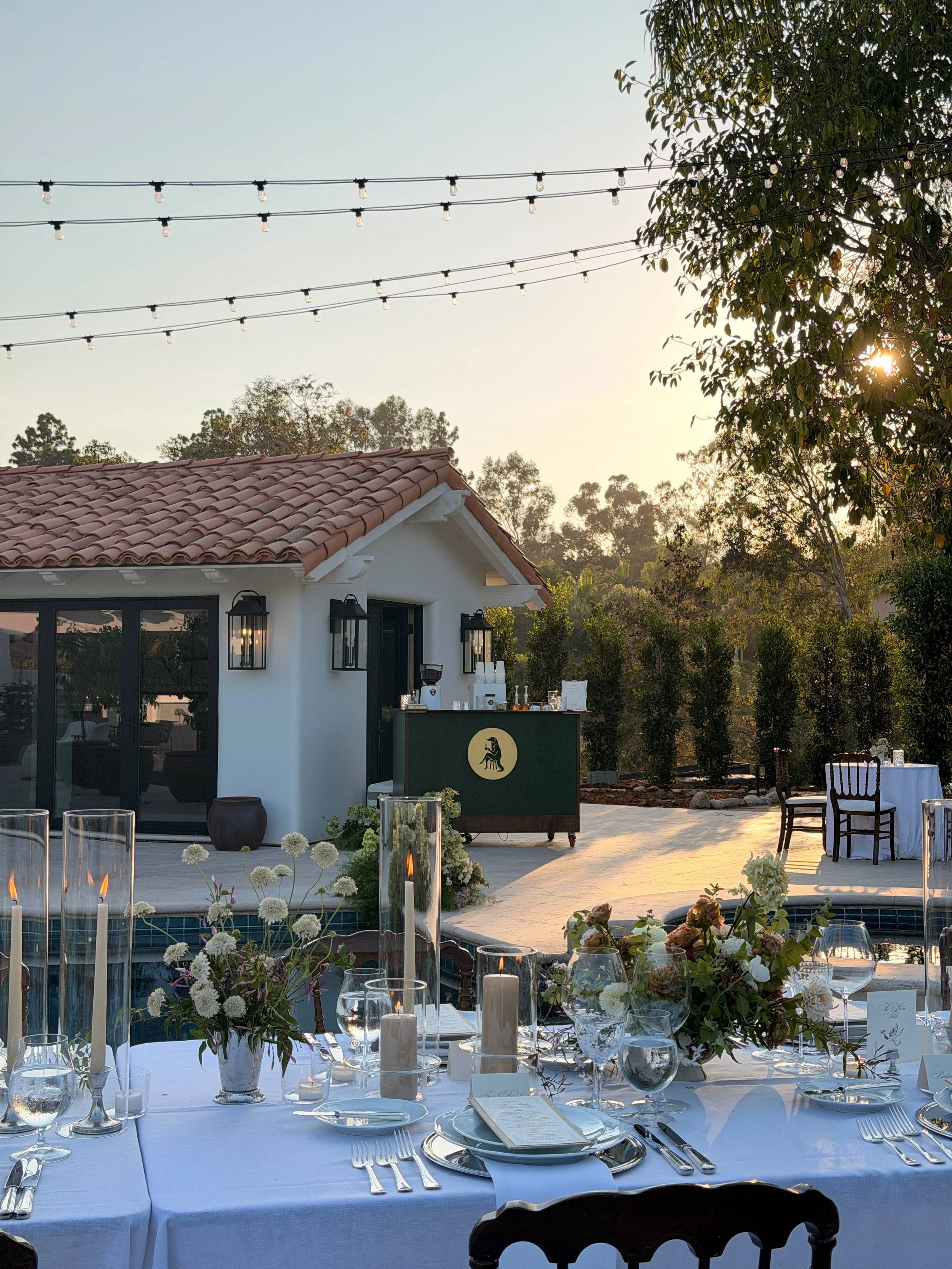 Elegant outdoor dining setup with a long white tablecloth, floral centerpieces, candles, and glassware, set for a formal event. In the background, a house with a clay tile roof, trees, and string lights are visible, with the sun setting behind the trees.