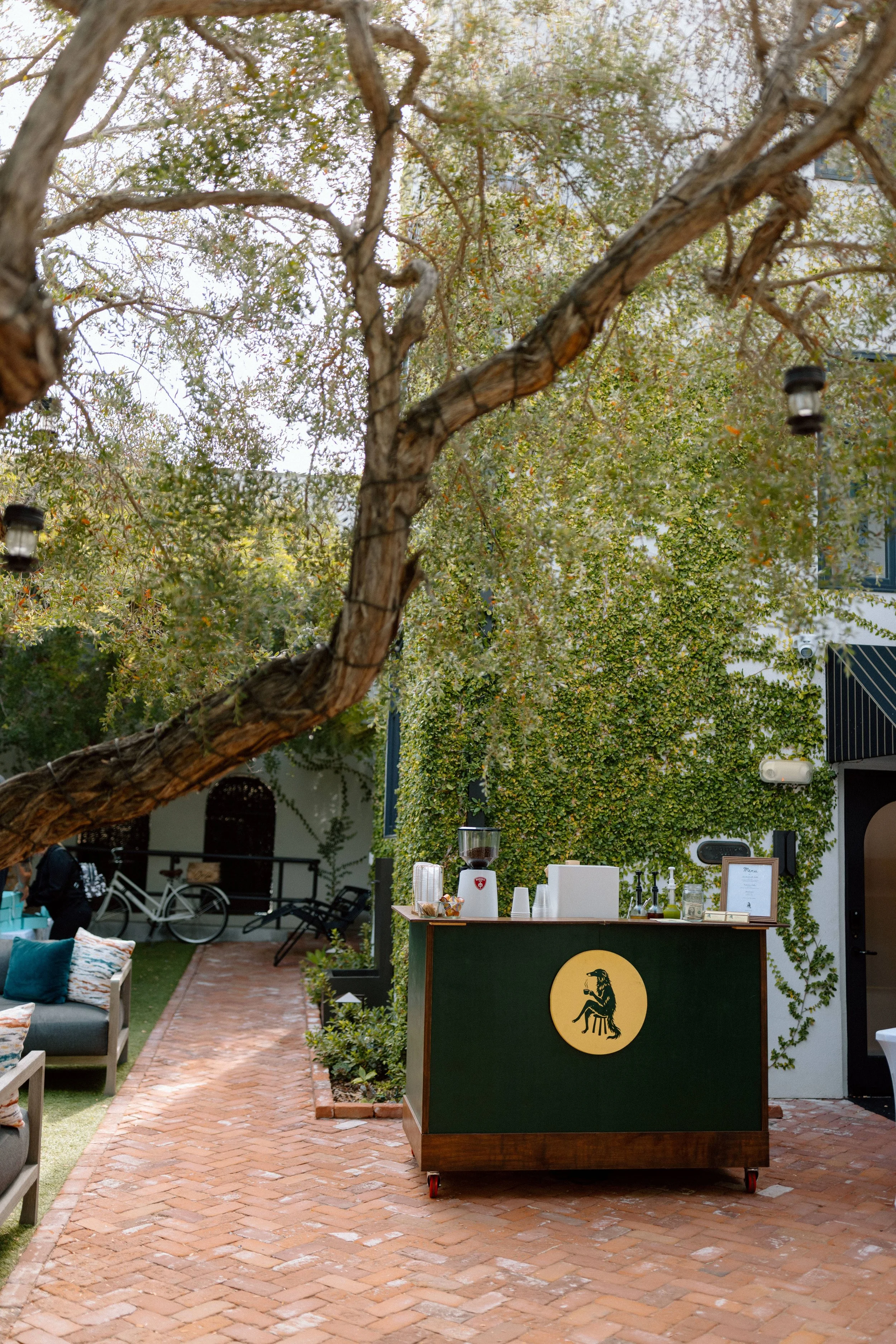 An outdoor cafe setup featuring a green serving cart with a yellow lion logo, surrounded by a brick pathway, trees, and seating areas with pillows. There are also some bicycles and a building in the background.