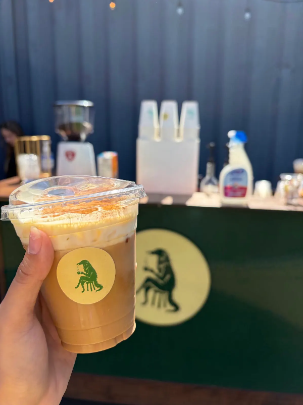 Person holding a plastic cup of iced coffee with whipped cream in front of a green and yellow stand with a lion logo.